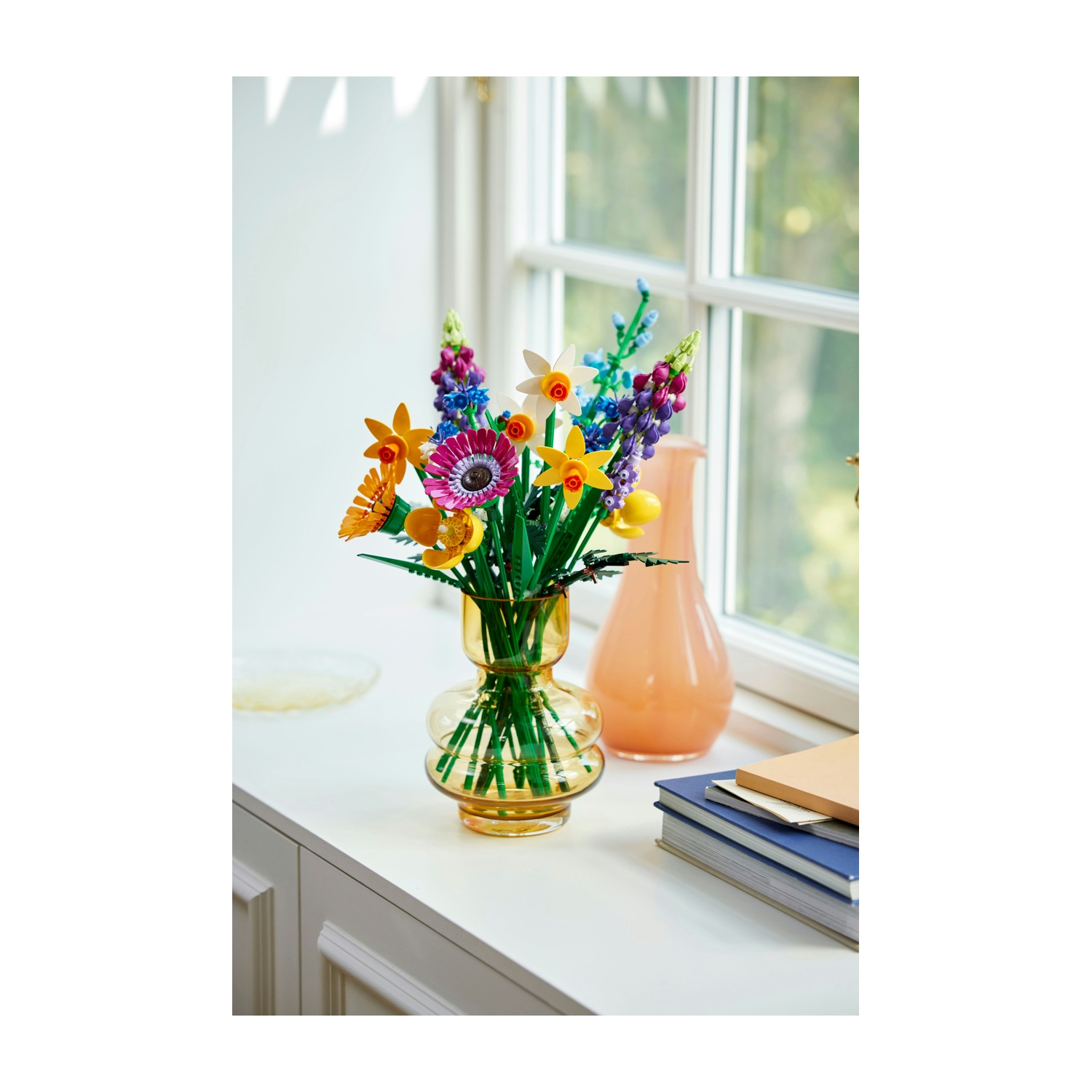 Un bouquet di fiori Lego colorati in un vaso giallo è su una mensola bianca. Un vaso color pesca e dei libri sono vicini, con una finestra sullo sfondo.