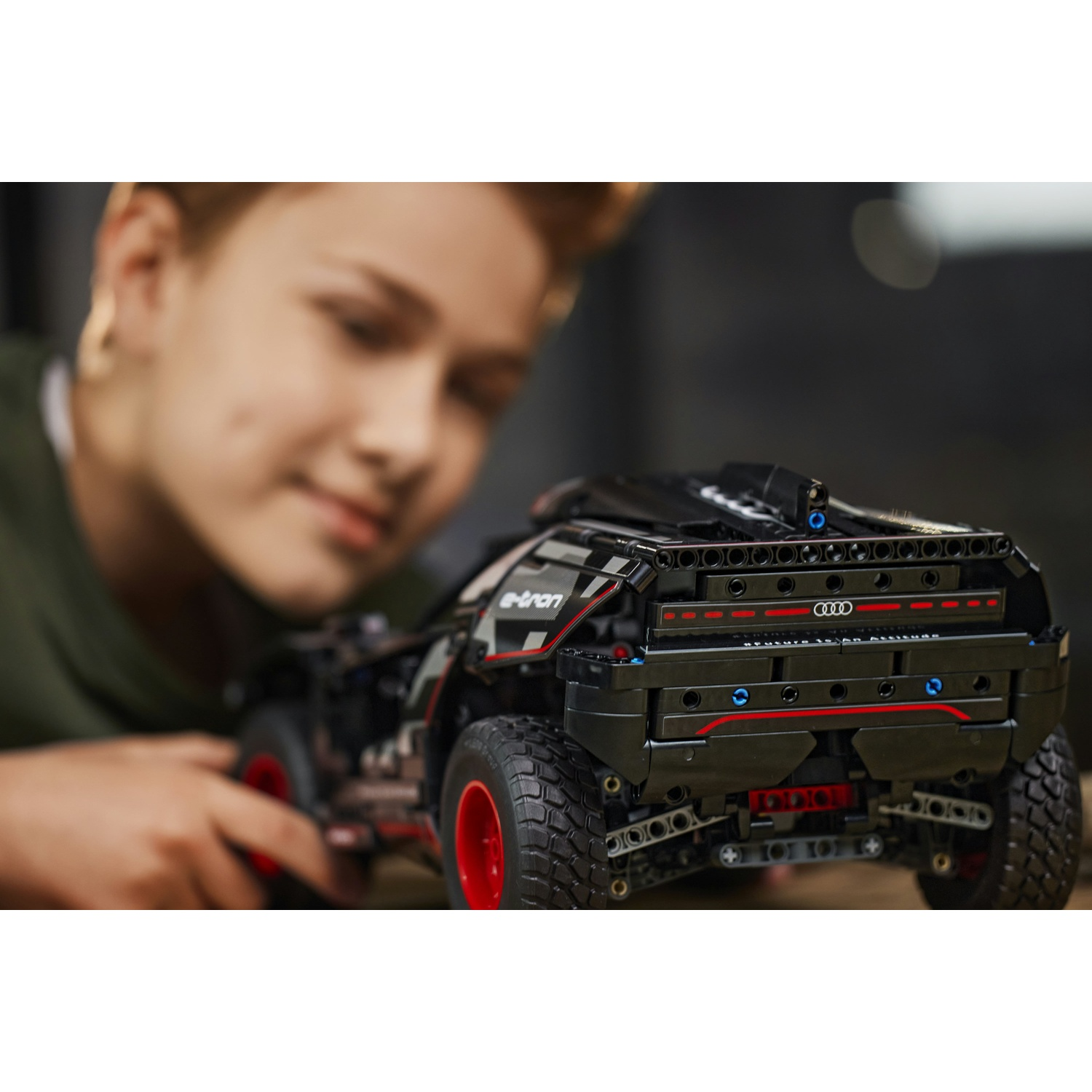 Una persona guarda un'auto Lego nera con ruote rosse. L'auto ha 'e-tron'. Lo sfondo è sfocato.