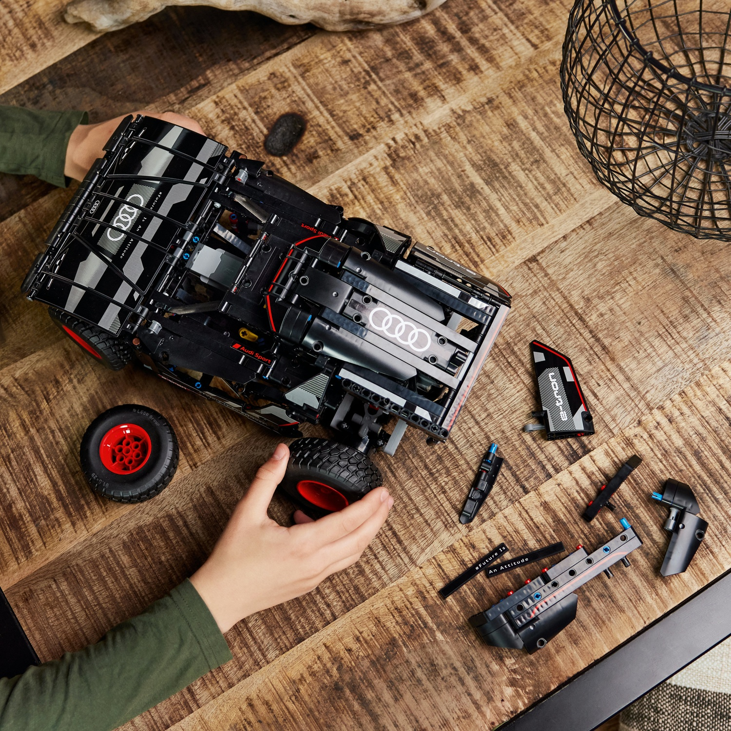Una persona assembla un modello di auto Audi nera. I pezzi sono su un tavolo di legno. L'auto ha ruote rosse. Un cestino di metallo sullo sfondo.