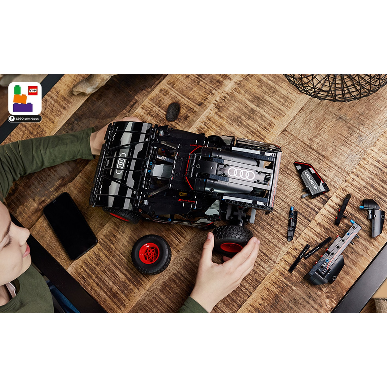 Una persona assembla un'auto Lego Audi nera su un tavolo di legno. Sul tavolo ci sono anche pezzi e un telefono. La persona indossa il verde.