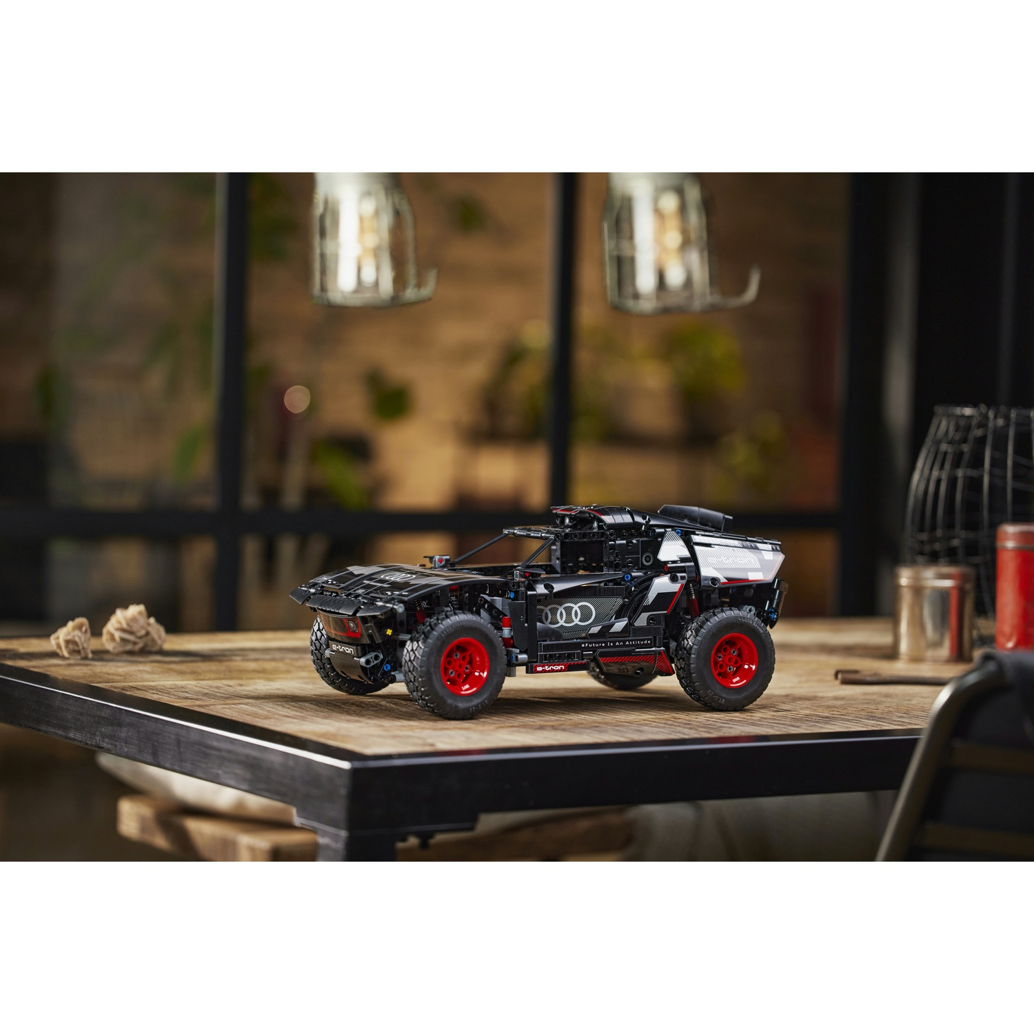 Un'auto da rally Audi Lego nera con ruote rosse è su un tavolo di legno. Lo sfondo comprende luci, piante e una sedia di metallo.
