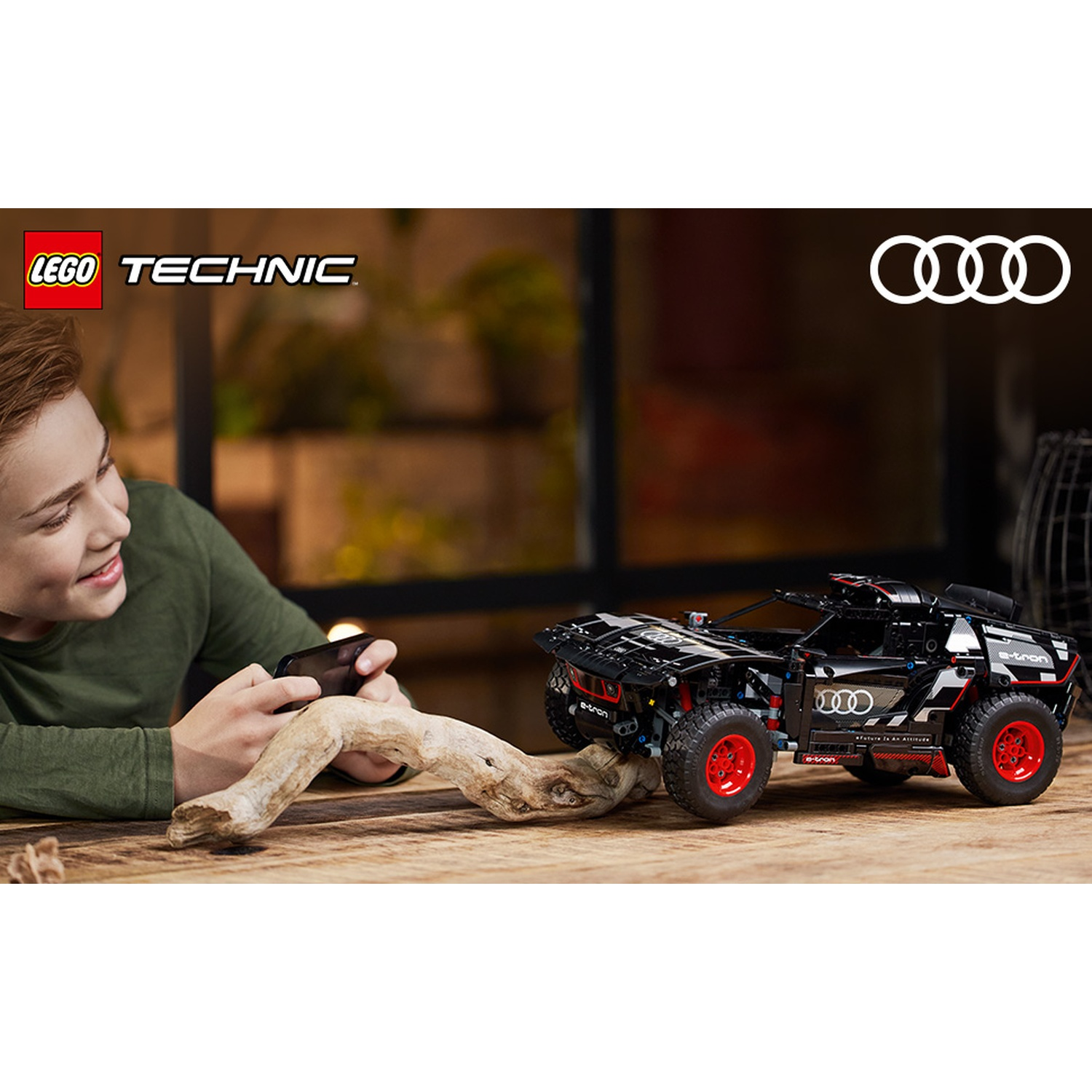 Un ragazzo sorride mentre usa un telefono. Un'auto Audi Lego nera con ruote rosse è su un tavolo di legno. I loghi Lego e Audi sono visibili.