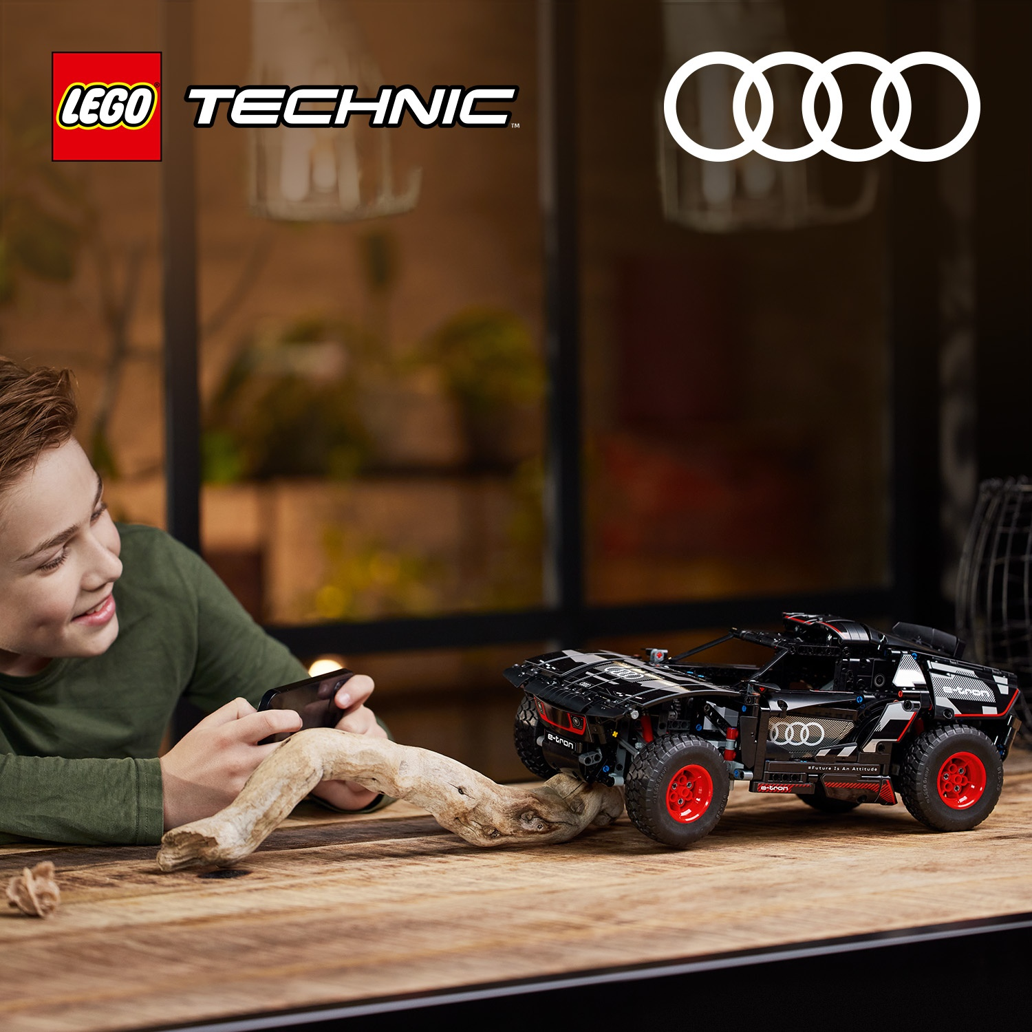 Un ragazzo sorride tenendo un telefono vicino a un modello di auto Lego Technic Audi e-tron su un tavolo di legno. Sfondo con logo Audi.