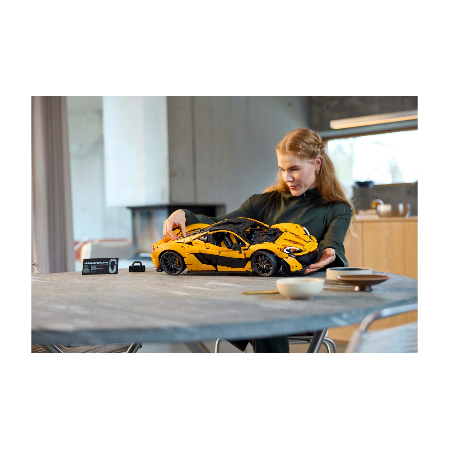 Una donna esamina una McLaren Lego gialla e nera su un tavolo grigio. Altri oggetti sono sul tavolo e un camino sullo sfondo.