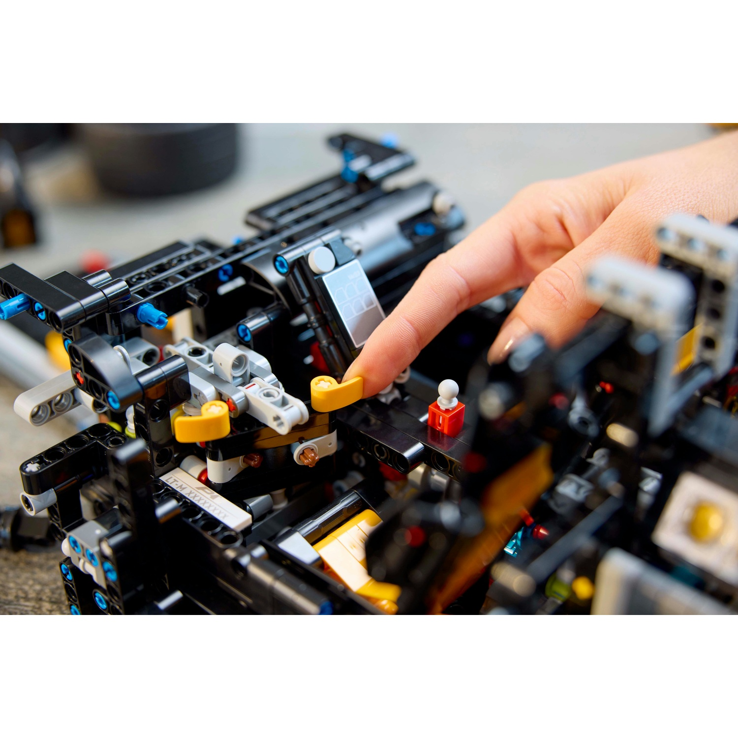 Una mano indica una complessa costruzione LEGO Technic nera. La costruzione presenta componenti gialli, blu e grigi. Una ruota è visibile sullo sfondo.
