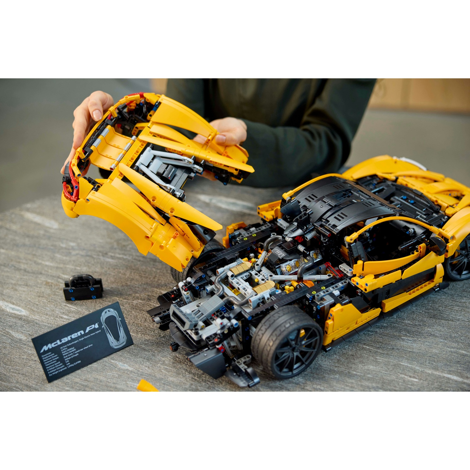 Una persona che assembla un modello Lego McLaren P1 giallo e nero. Le parti vengono tenute e posizionate su un tavolo con una scheda dati.