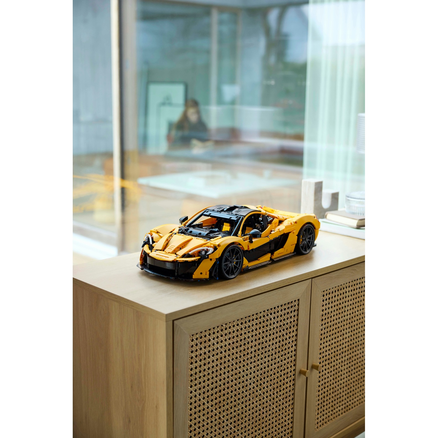 Un modello di auto Lego McLaren P1 giallo e nero è su un mobile di legno. Sullo sfondo si vedono una finestra e una persona.