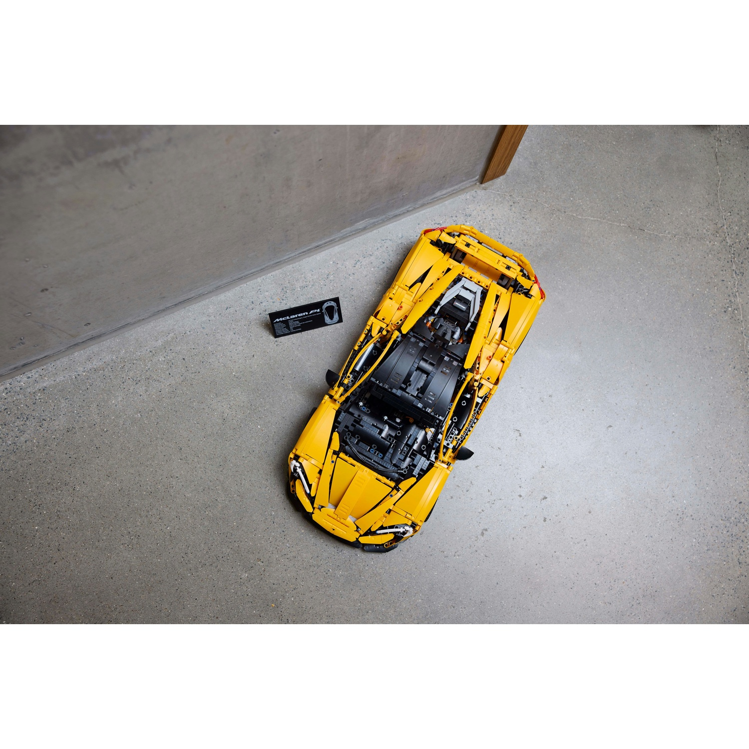 Un'auto McLaren Lego gialla è su un pavimento di cemento grigio. Una scatola nera e un muro sono a sinistra. Vista dall'alto.
