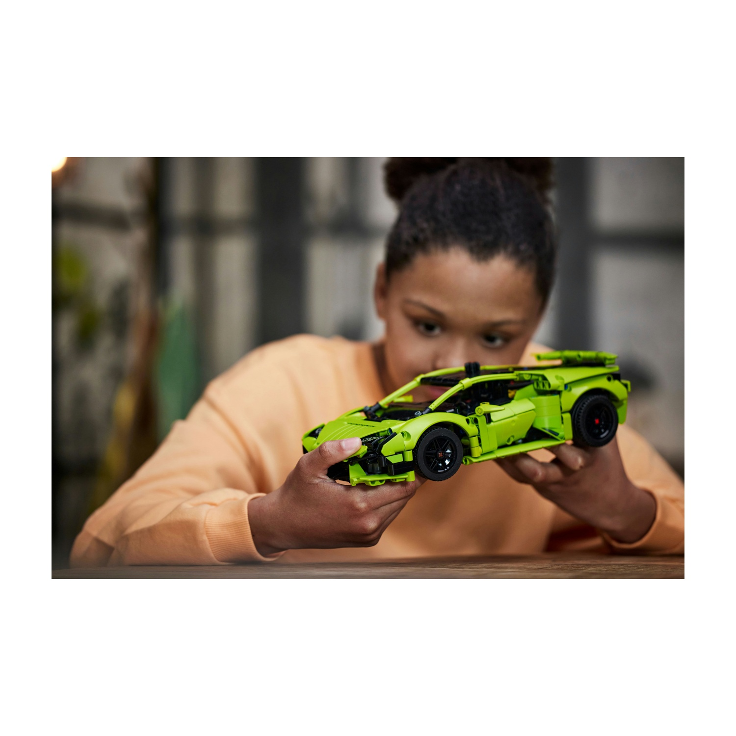 Una persona tiene in mano un'auto giocattolo verde lime, concentrandosi su di essa. Indossa un maglione arancione. L'auto ha ruote nere.
