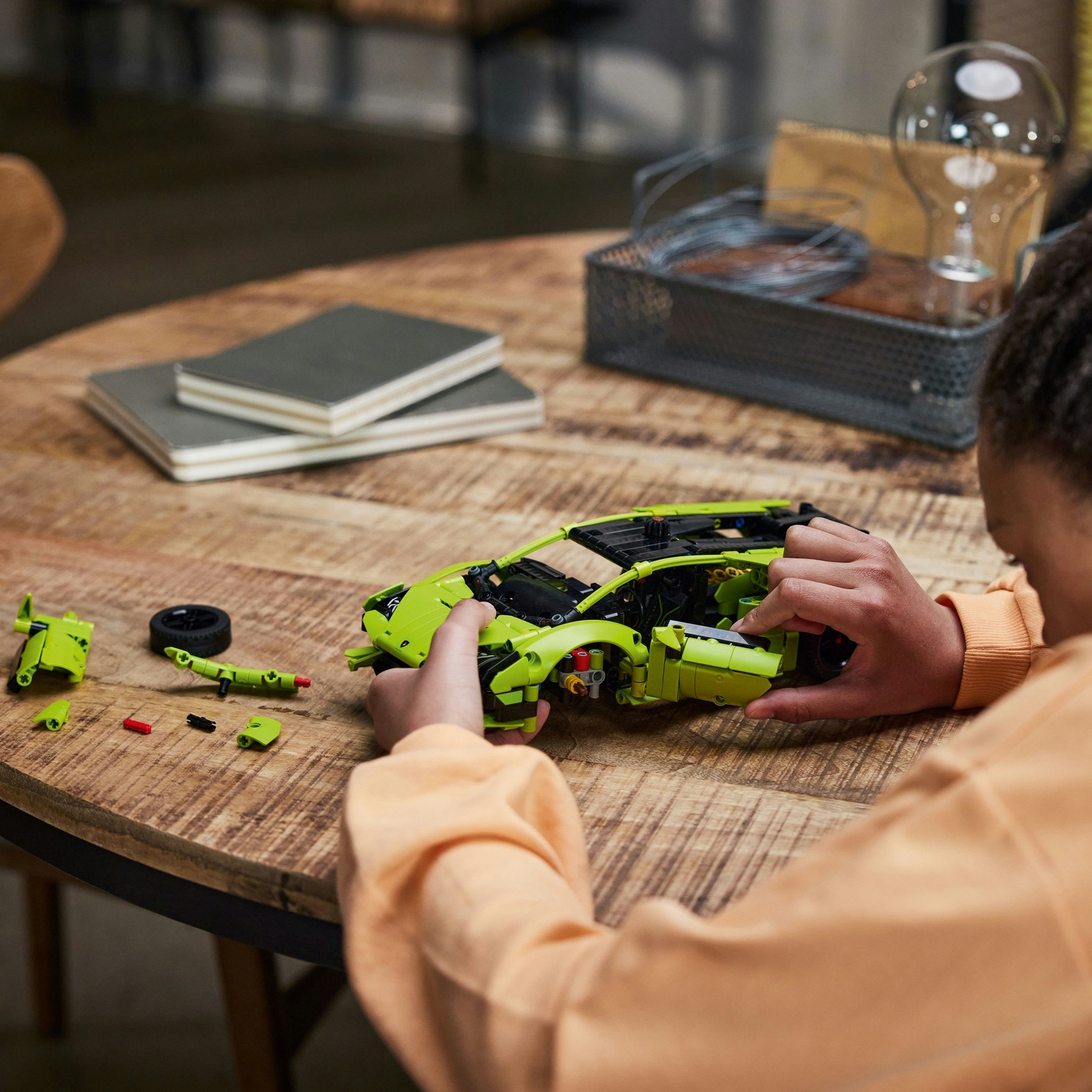Una persona assembla un'auto giocattolo verde su un tavolo di legno. I pezzi sono sparsi. Una lampadina e dei quaderni sono sullo sfondo.