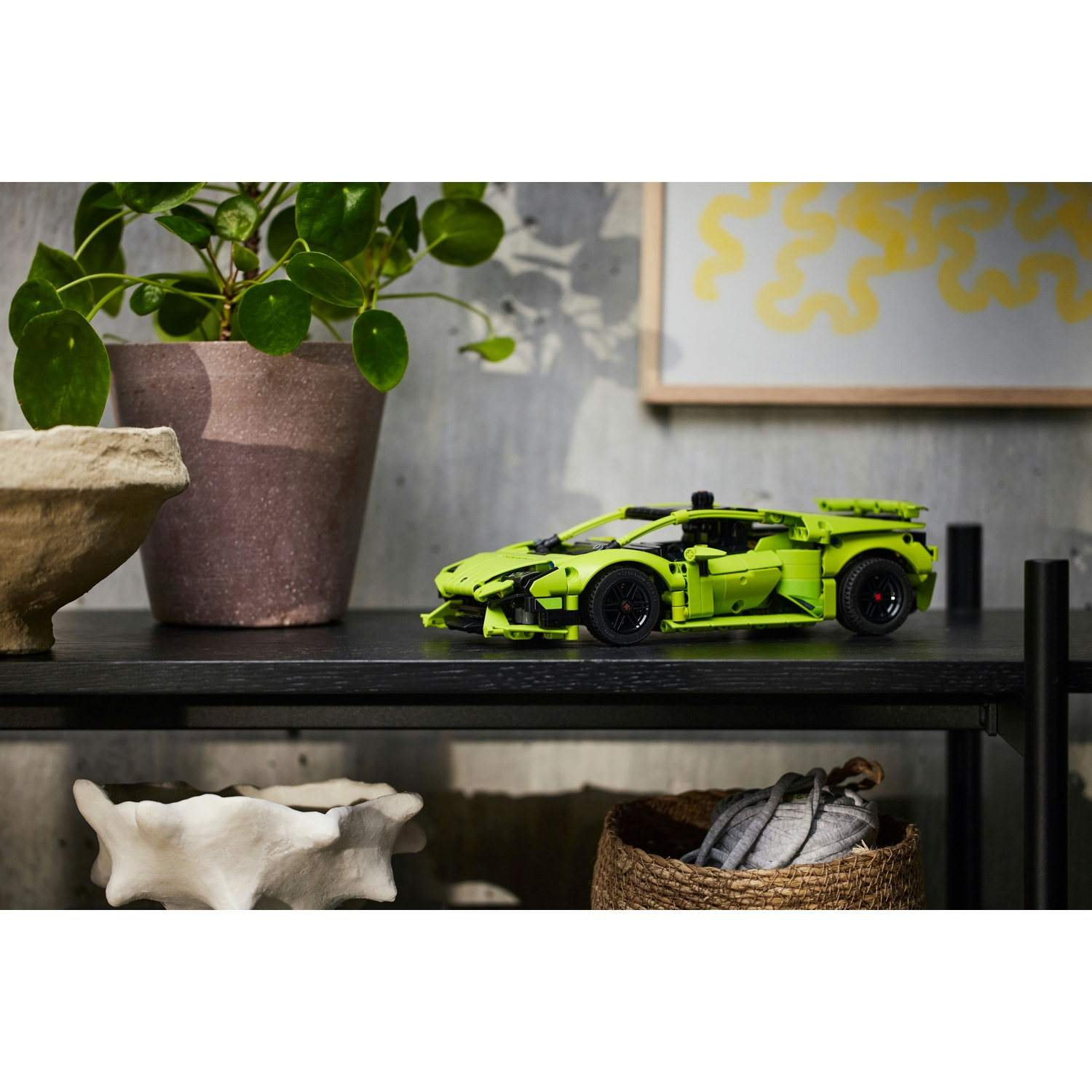 Un'auto giocattolo verde lime è su una mensola nera con piante e una ciotola decorativa. Un dipinto con scarabocchi gialli è appeso sullo sfondo.