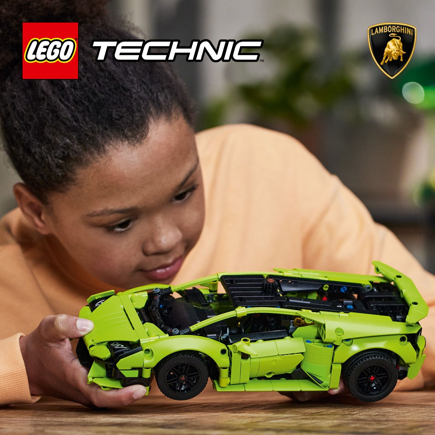 Un bambino tiene in mano una Lamborghini Lego verde. I loghi Lego e Lamborghini sono visibili. Il bambino indossa un maglione arancione e guarda l'auto.