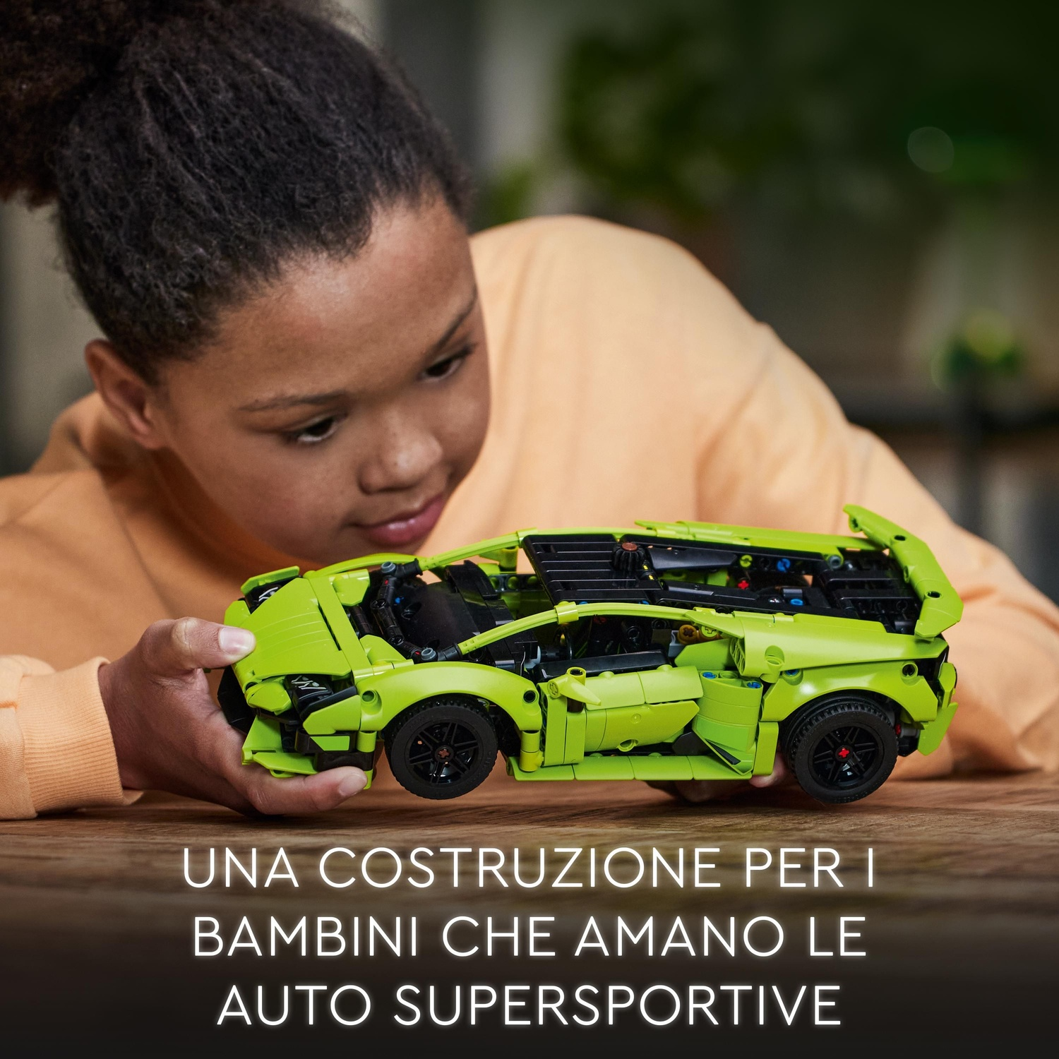 Un giovane tiene in mano un'auto sportiva giocattolo verde lime. L'auto è su un tavolo di legno. Testo in italiano in basso.