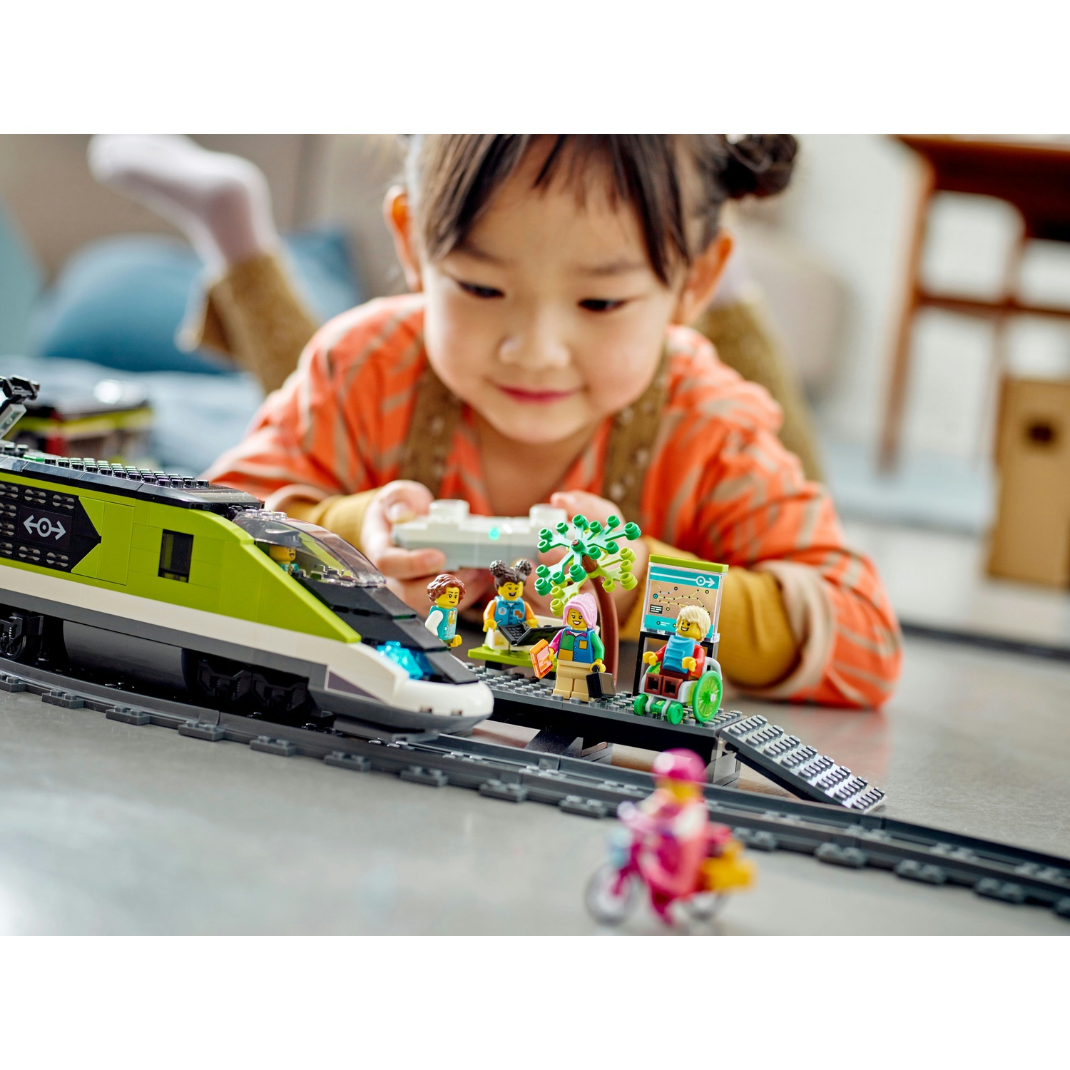 Una bambina gioca con un trenino Lego. Un treno verde e bianco sui binari. Ci sono personaggi Lego e una rampa.