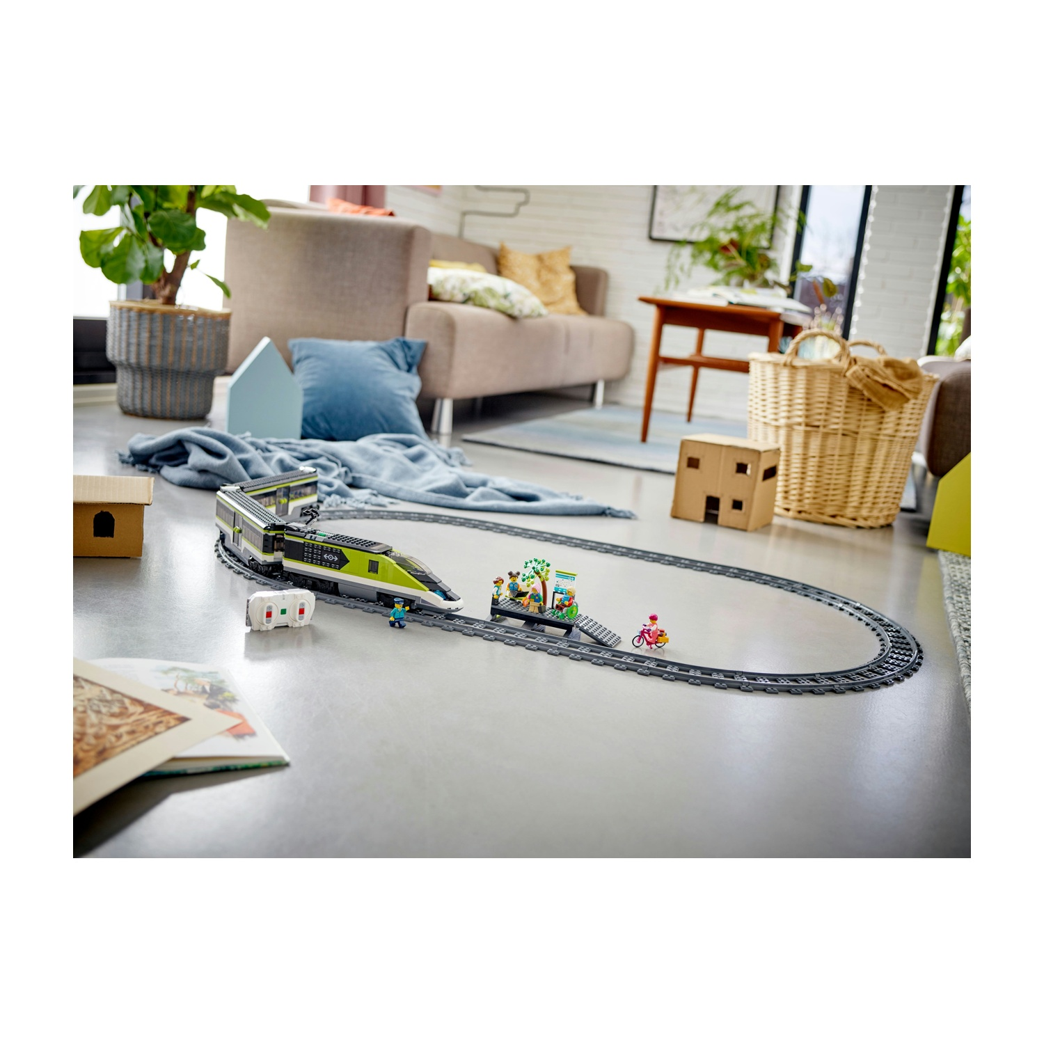 Un trenino giocattolo sui binari in una stanza. Un treno verde e grigio, con una piccola stazione. Un bambino va in bicicletta.