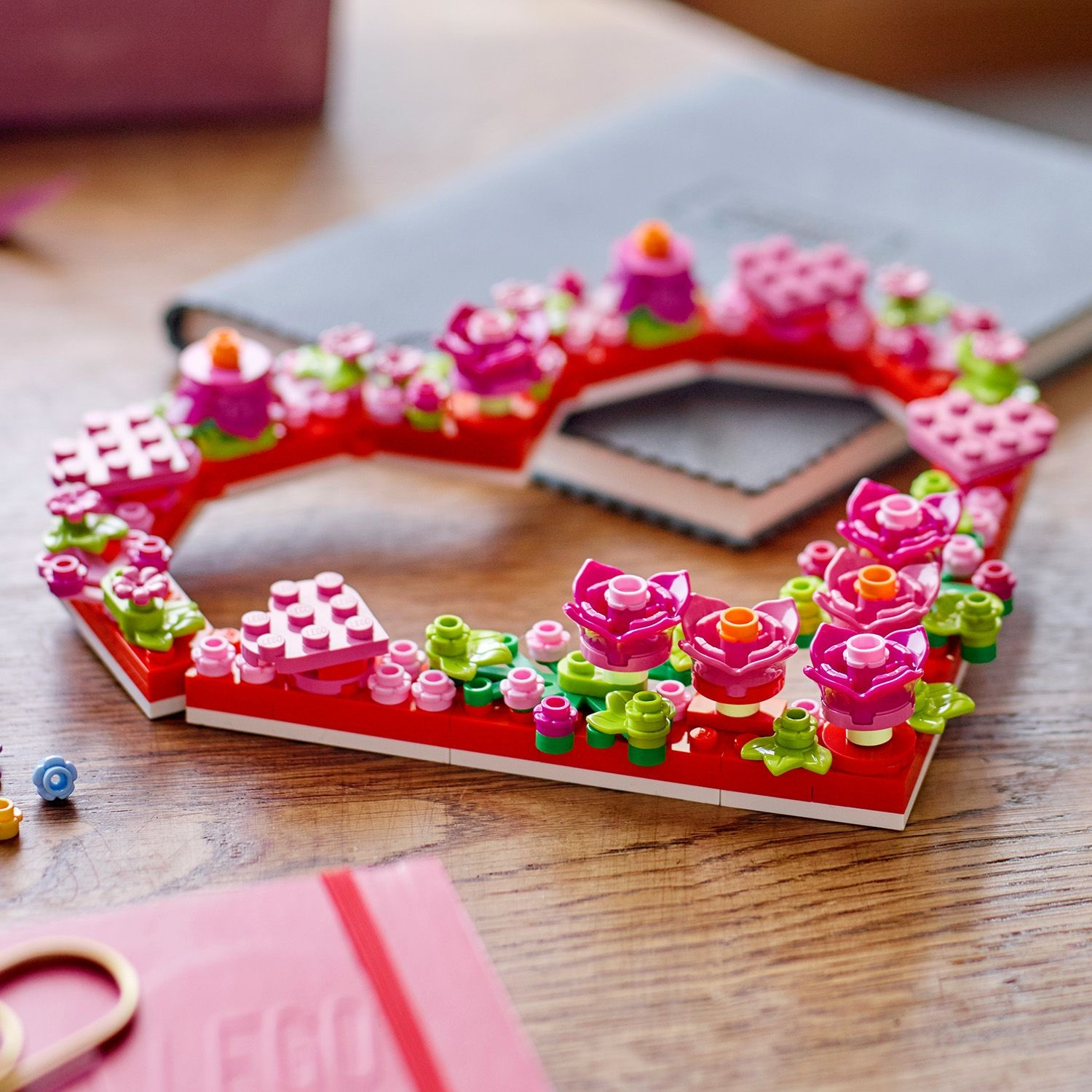 Una cornice floreale Lego con fiori rosa e foglie verdi. Si trova su un tavolo di legno, con un taccuino sullo sfondo.