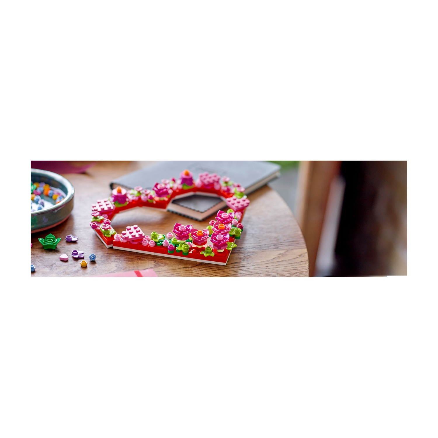 Un tavolo di legno con una cornice Lego a forma di cuore decorata con fiori. Un quaderno, una ciotola con perline colorate e piccoli pezzi Lego sono vicini.