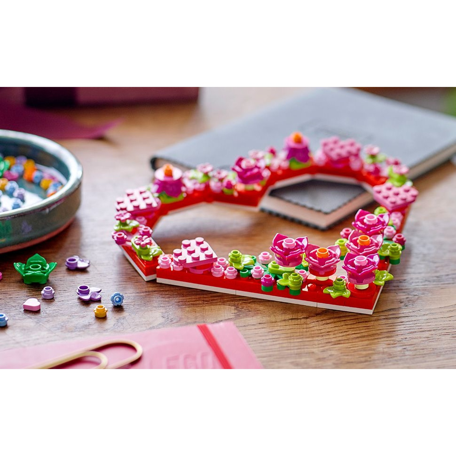 Una creazione Lego a forma di cuore con fiori e candele rosa è su un tavolo di legno, insieme a pezzi Lego sparsi e un taccuino.