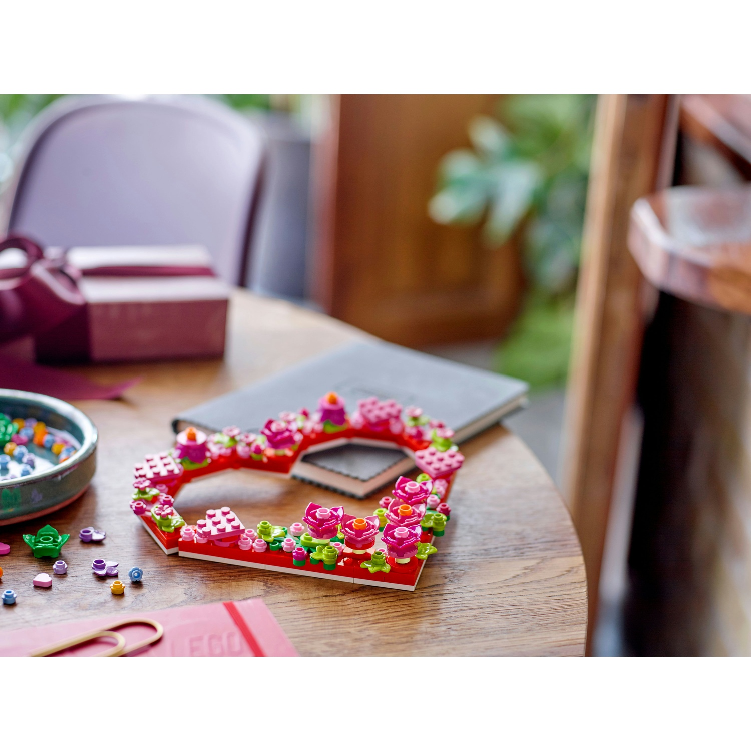 Una decorazione Lego a forma di cuore con fiori rosa. È su un tavolo di legno, con un taccuino, una scatola regalo e pezzi Lego sparsi.