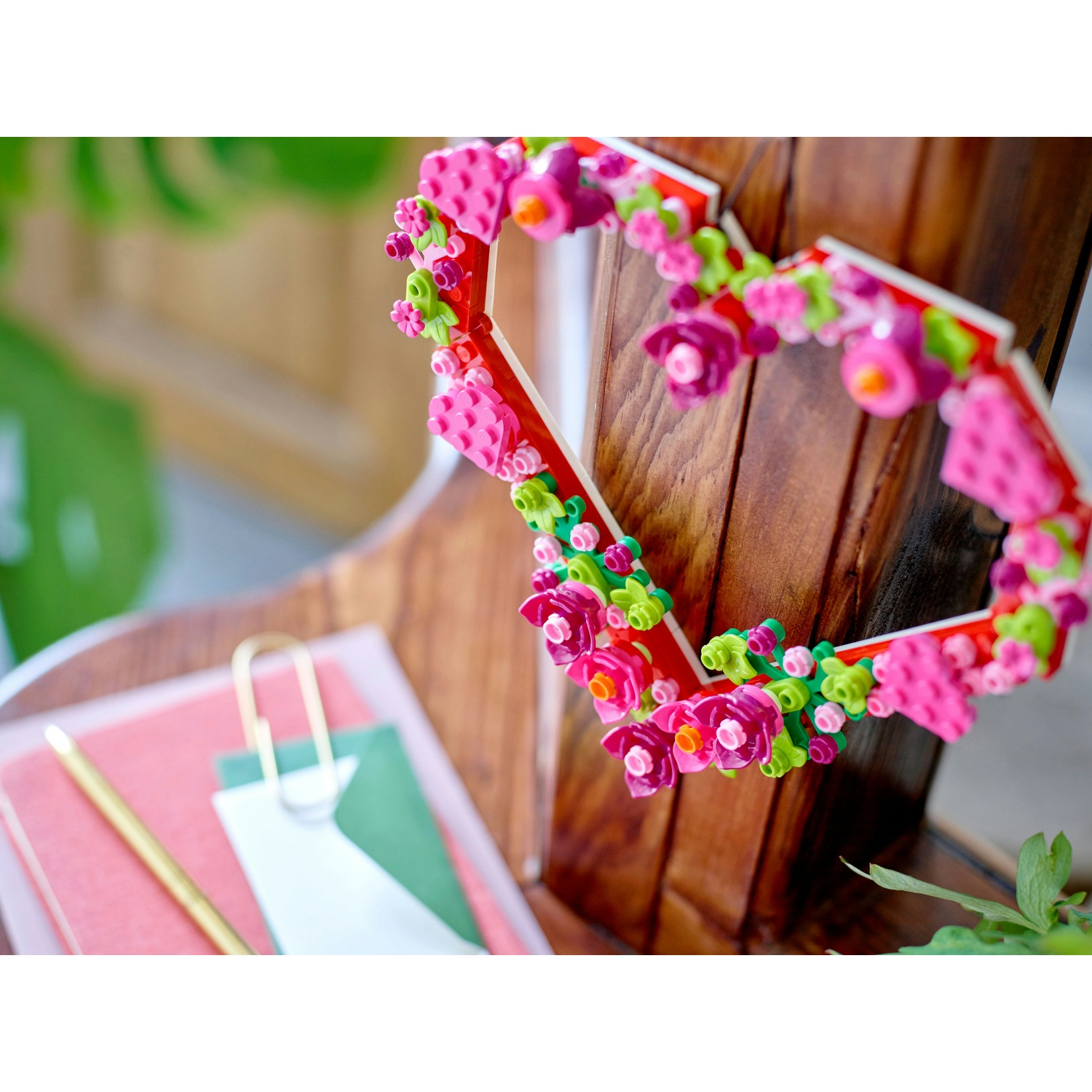 Una cornice a forma di cuore in legno decorata con fiori rosa. Poggia su una superficie di legno, con un blocco note e una penna in primo piano.