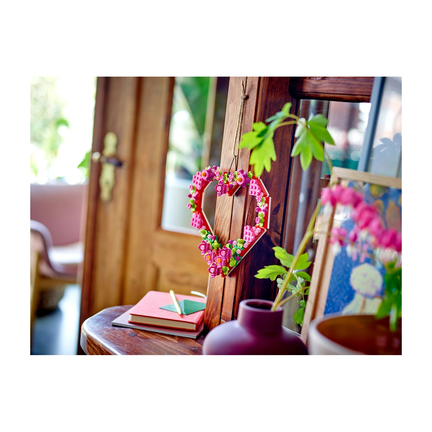Un cuore di legno decorato con fiori è appeso a una struttura in legno. Un libro rosso e carta verde sono su un tavolo di legno.
