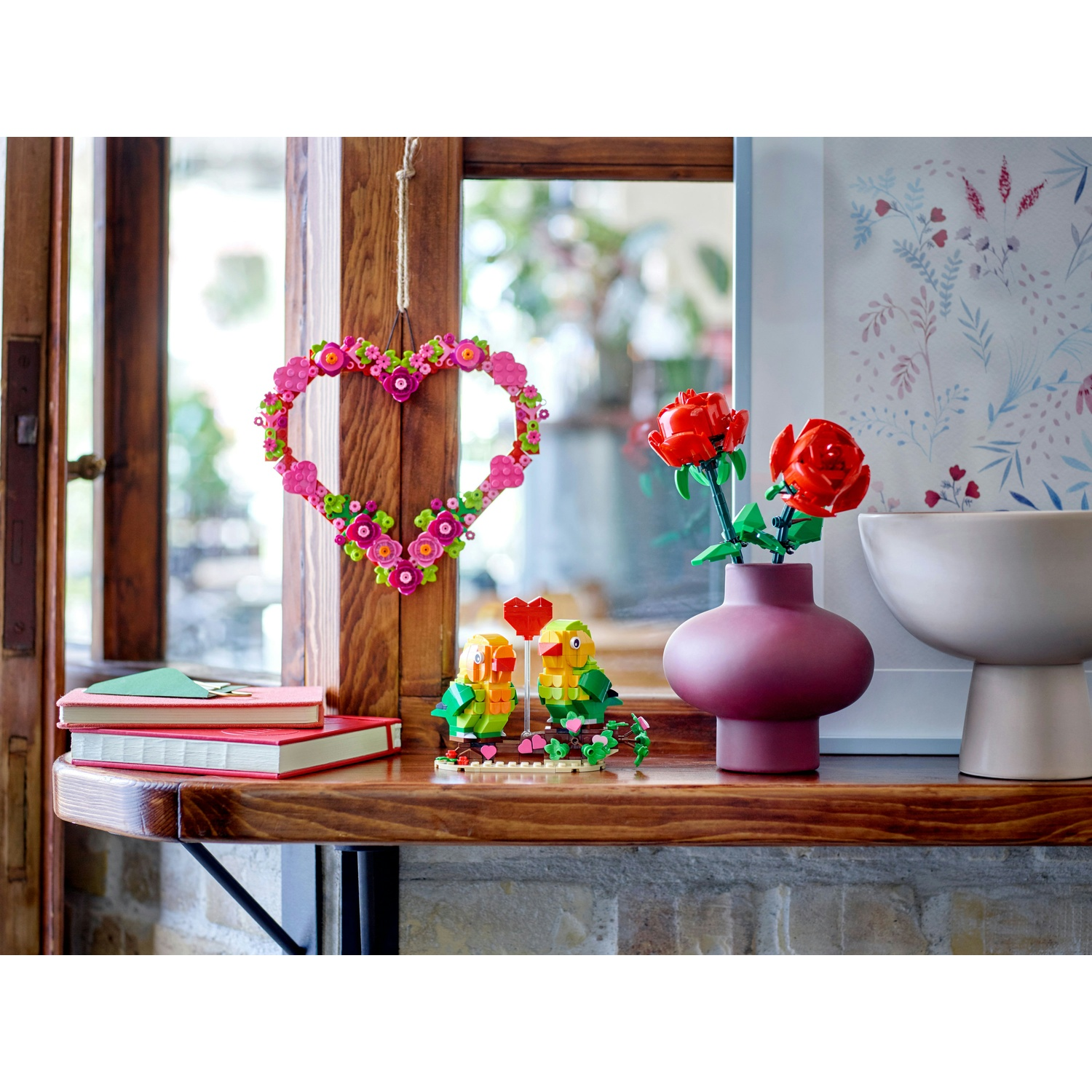 Una mensola di legno mostra una ghirlanda floreale a forma di cuore, uccelli Lego, rose rosse in un vaso e libri, il tutto in un ambiente luminoso.