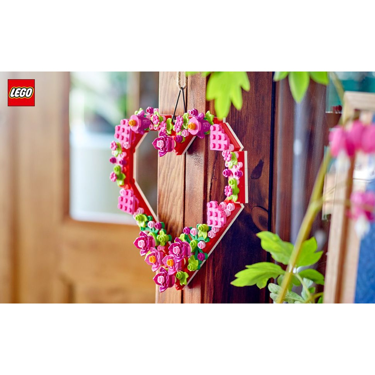 Una ghirlanda Lego a forma di cuore con fiori rosa è appesa a una superficie di legno. Un logo Lego è nell'angolo in alto a sinistra.