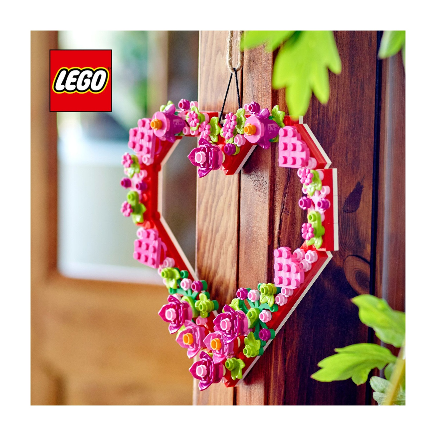 Una ghirlanda Lego a forma di cuore con fiori rosa, cornice rossa e foglie verdi, appesa a una porta di legno. Logo Lego a sinistra.