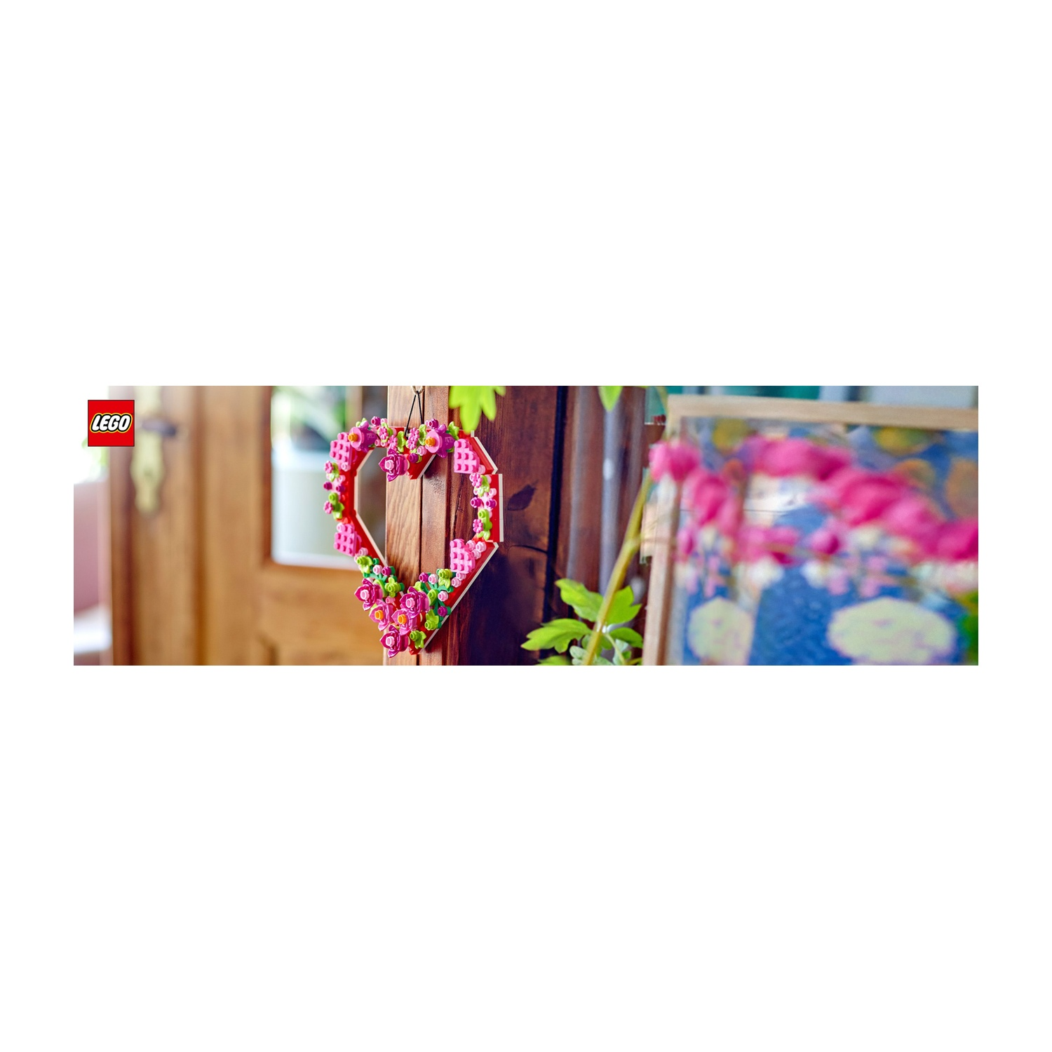 Una decorazione Lego a forma di cuore con fiori rosa e verdi è appesa a una superficie di legno. Un dipinto sfocato sullo sfondo.