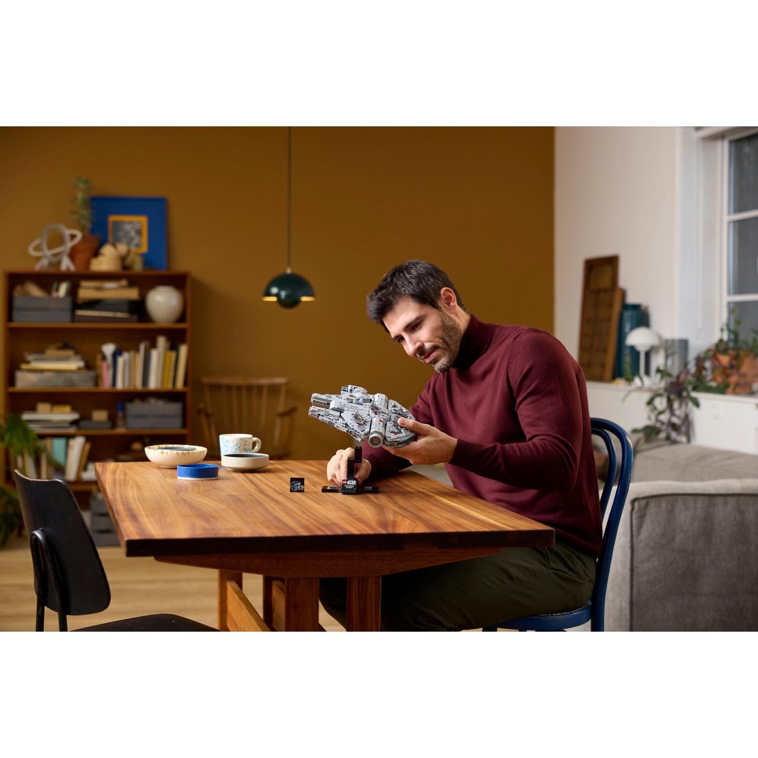 Un uomo con un maglione bordeaux è seduto a un tavolo di legno, tenendo in mano un modellino di astronave. Librerie e un divano sullo sfondo.