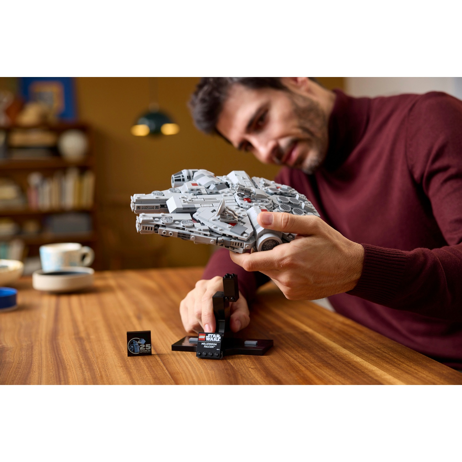Un uomo tiene in mano un Millennium Falcon Lego. Lo sta guardando. Un piccolo espositore e un supporto sono di fronte a lui su un tavolo di legno.