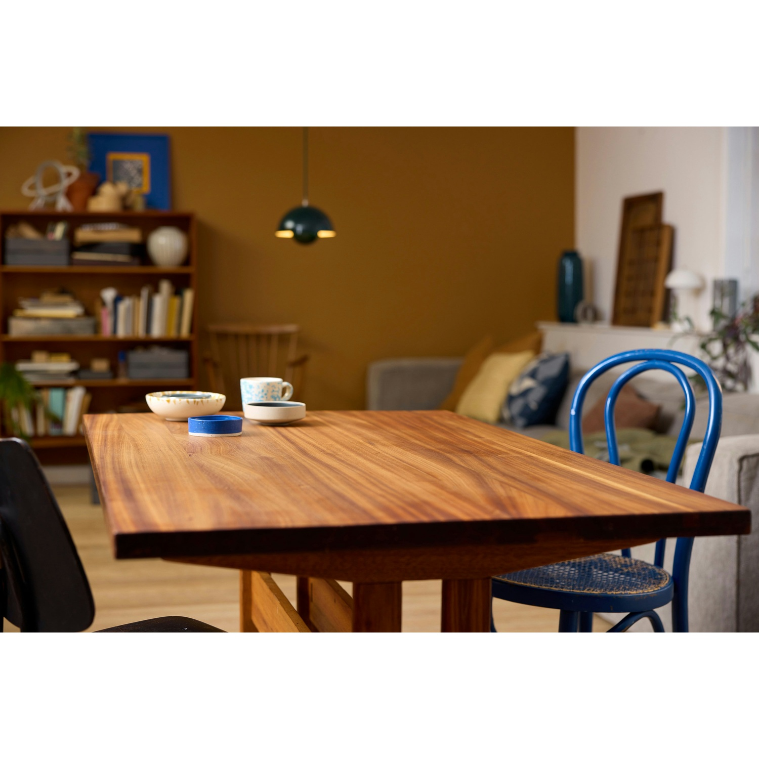 Tavolo in legno con piatti. Sedia blu a destra. Libreria e divano sullo sfondo. La lampada a soffitto è sopra.