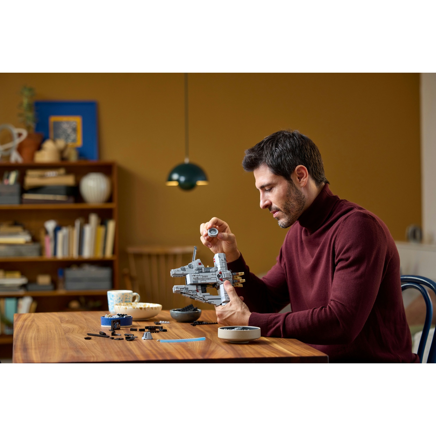Un uomo con un maglione bordeaux assembla un modello Lego su un tavolo di legno. La stanza ha una libreria e una lampada a sospensione.