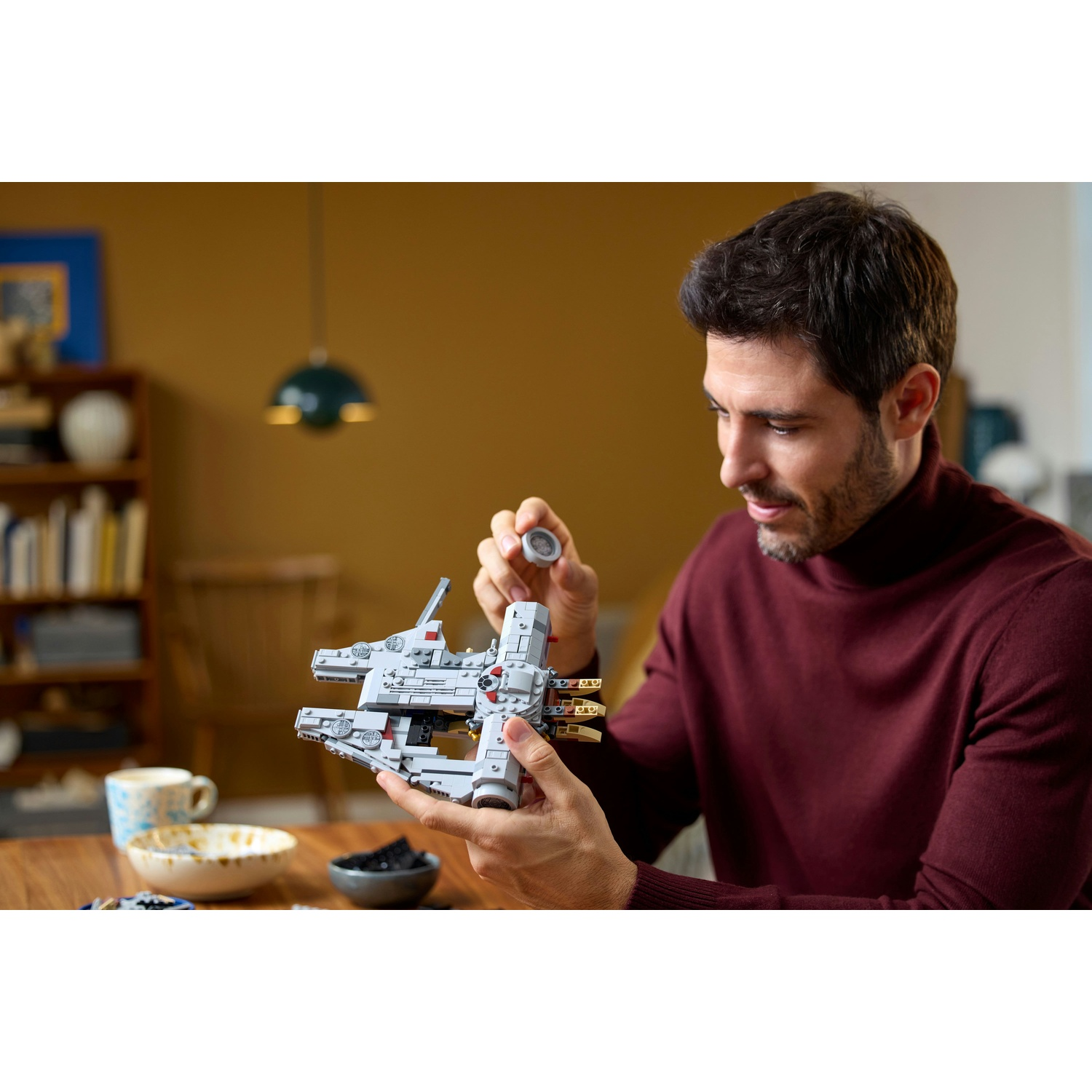 Un uomo con un maglione bordeaux assembla un'astronave Lego. Tiene un piccolo pezzo grigio sopra il modello su un tavolo di legno.