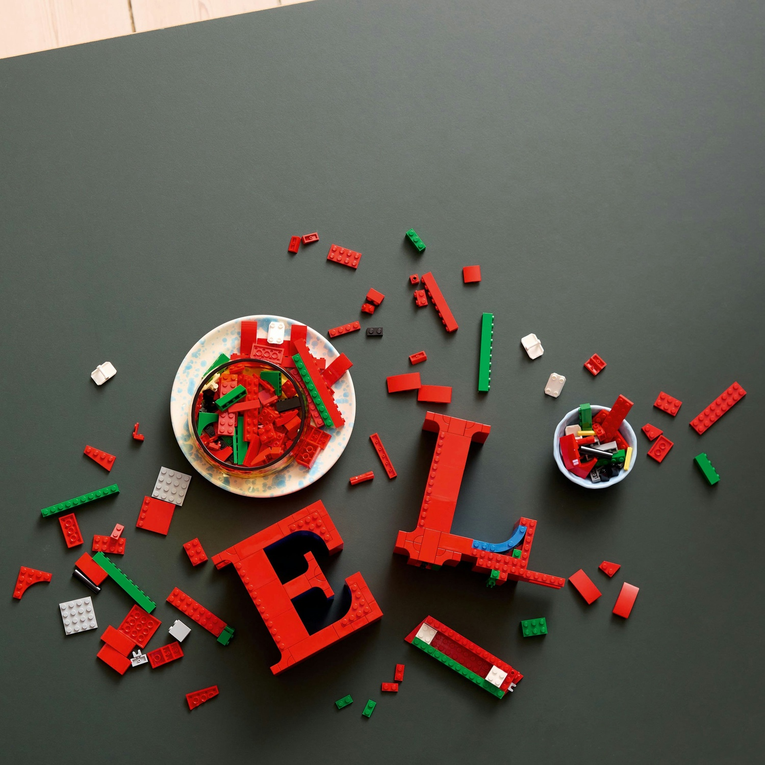 Lettere Lego rosse 'E' e 'L' con blocchi sparsi su una superficie grigio scuro. Una ciotola e un piatto contengono altri blocchi.