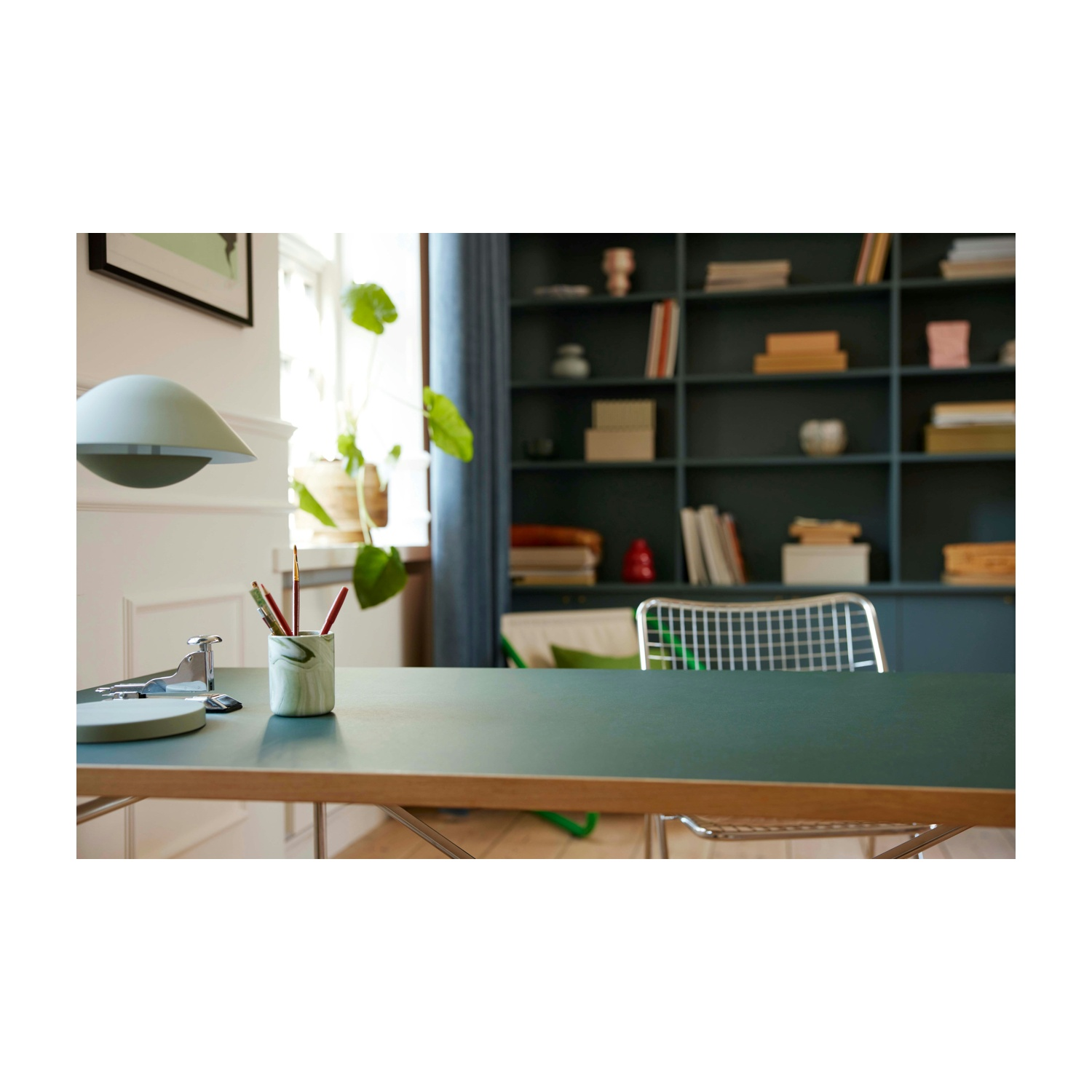 Una scrivania con materiali artistici. Una lampada è appesa sopra. Una libreria e una finestra sullo sfondo.
