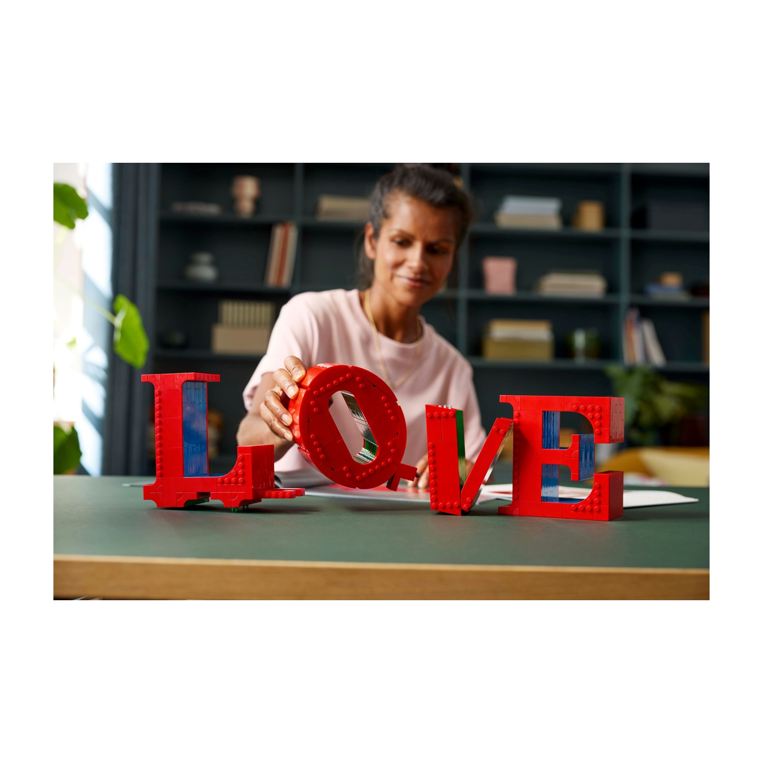 Una donna sistema lettere LEGO rosse che compongono 'LOVE'. Lo sfondo include una libreria e un tavolo verde.