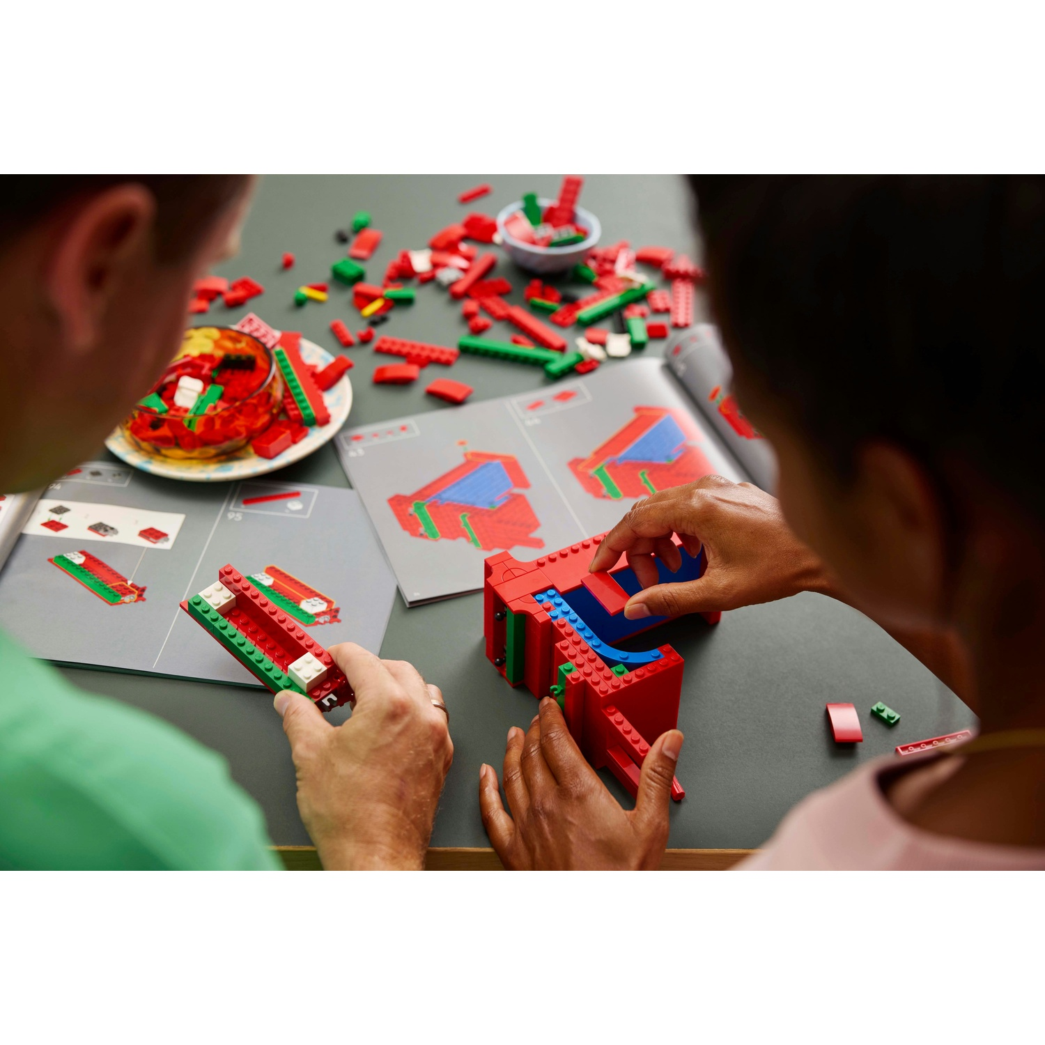 Due persone che costruiscono con i Lego. Vengono assemblati mattoncini rossi, verdi e bianchi. Istruzioni e snack sono sul tavolo.