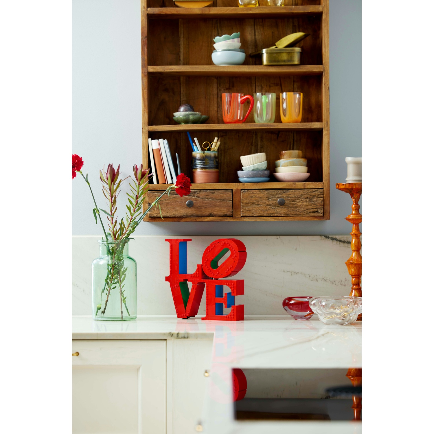 Una scena di cucina con una mensola di legno piena di oggetti, un'insegna 'LOVE', fiori in un vaso e un piano di lavoro con ciotole.