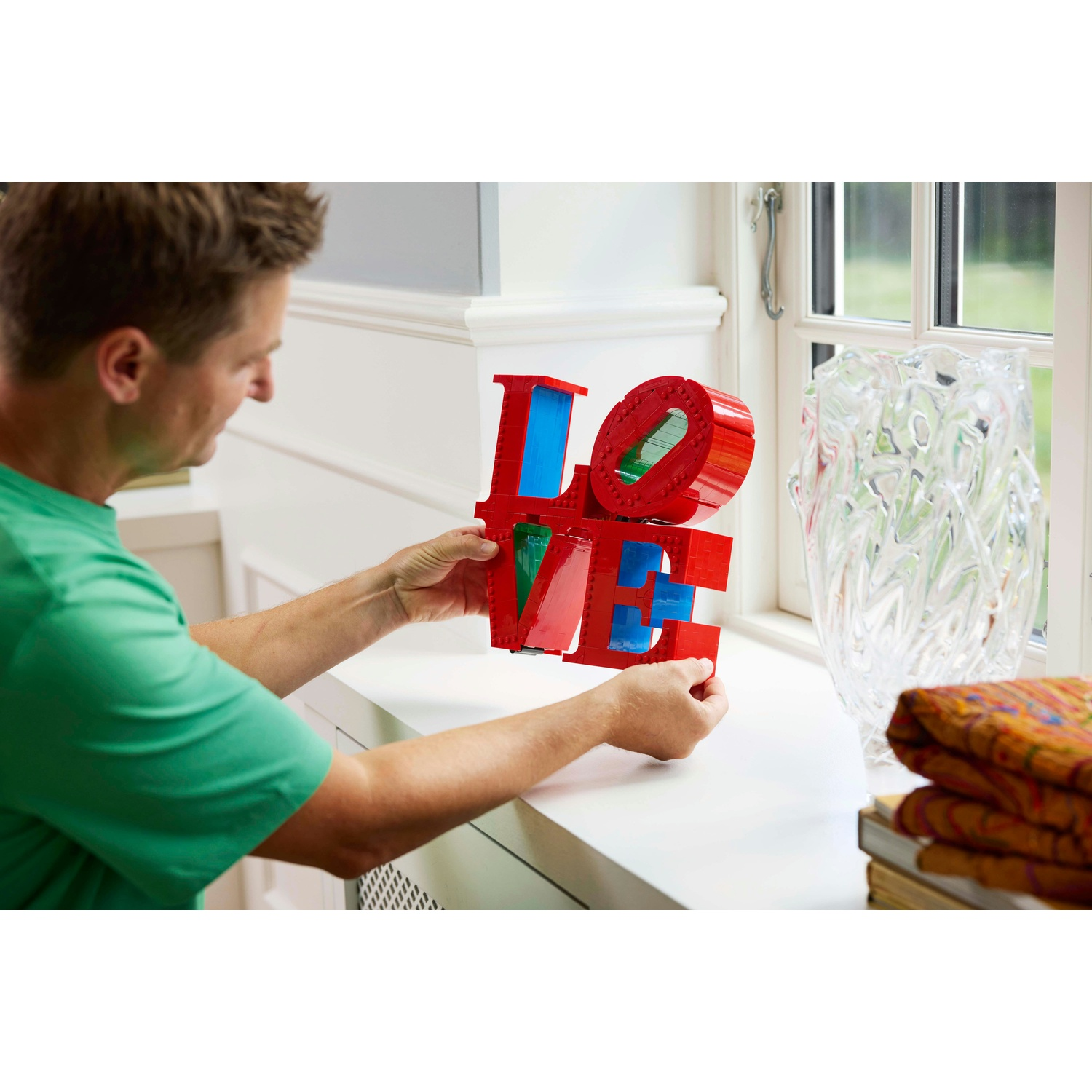 Un uomo tiene in mano un cartello Lego rosso 'LOVE' con accenti blu e verdi, appoggiandolo su un davanzale bianco. Un vaso e tessuti sullo sfondo.