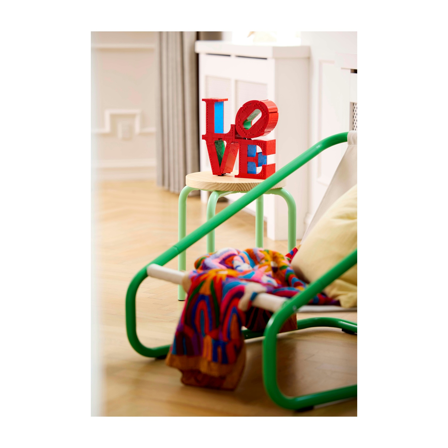 Una stanza con lettere 'LOVE' su uno sgabello e una sedia verde con una coperta. La scena è luminosa e ariosa, con pavimento in legno.