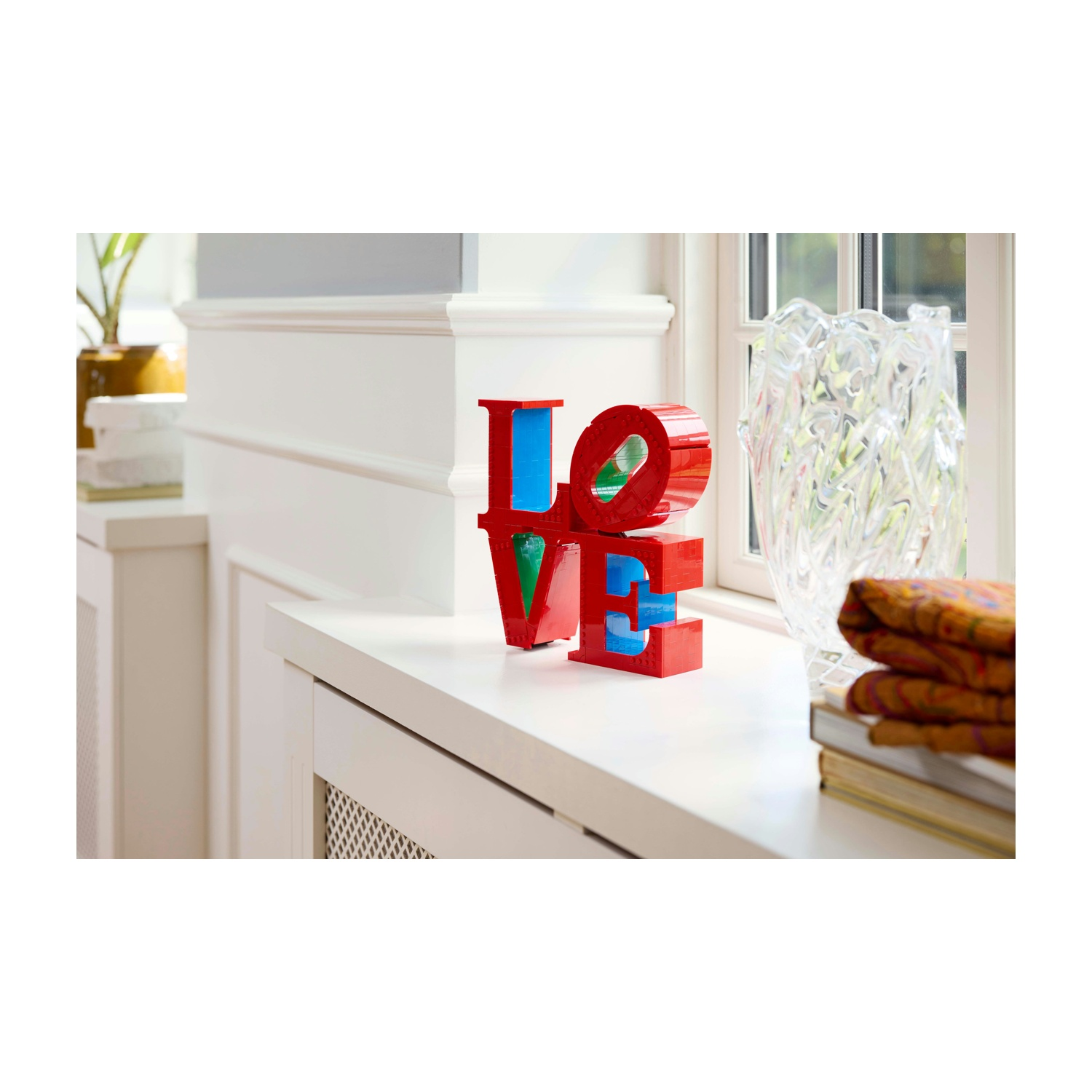 Una scultura "LOVE" rossa, blu e verde è su una superficie bianca. Un vaso trasparente, libri e tessuto sono a destra vicino a una finestra.