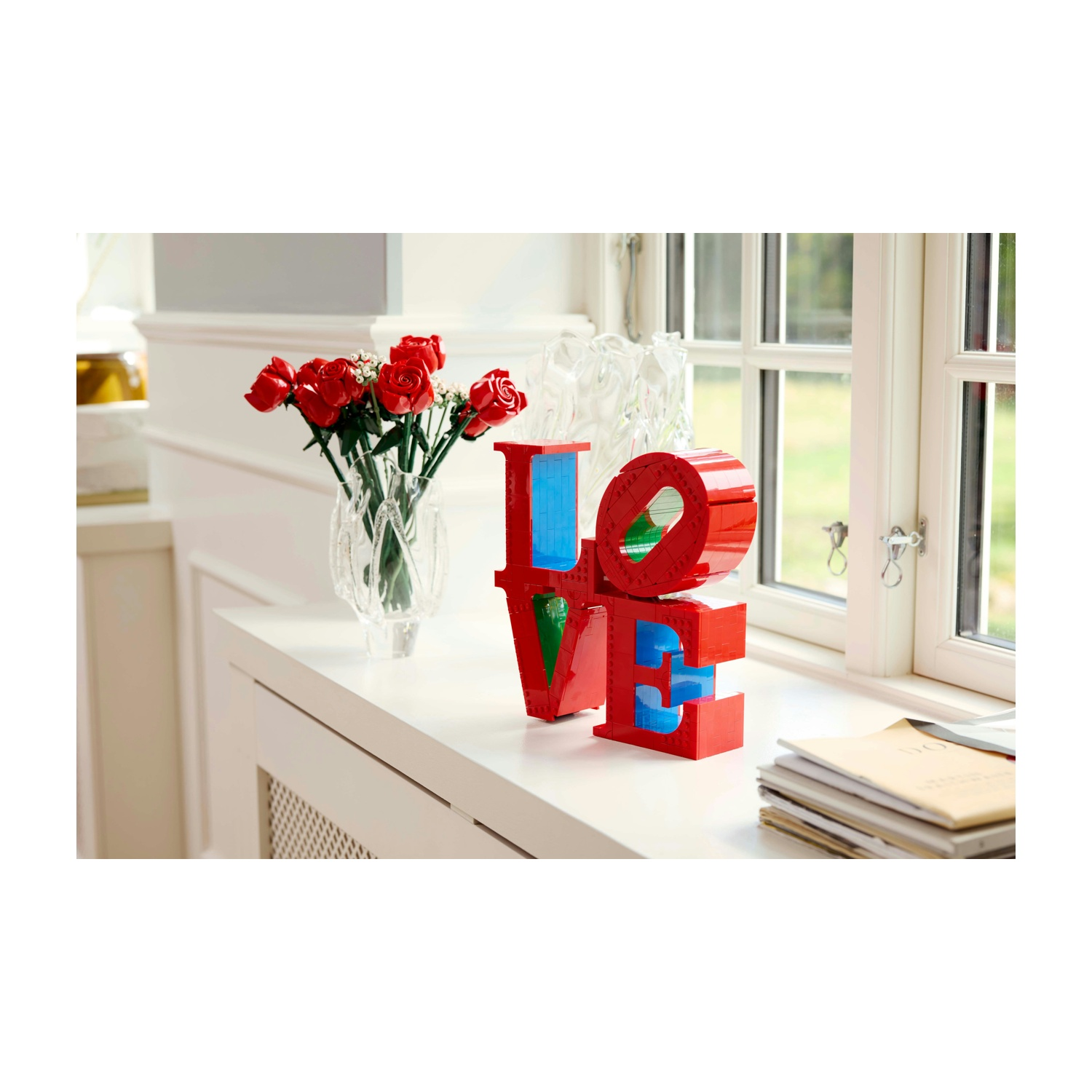 Scultura rossa 'LOVE' con accenti blu, un vaso di rose rosse e una finestra con vista sul verde, su una superficie bianca.