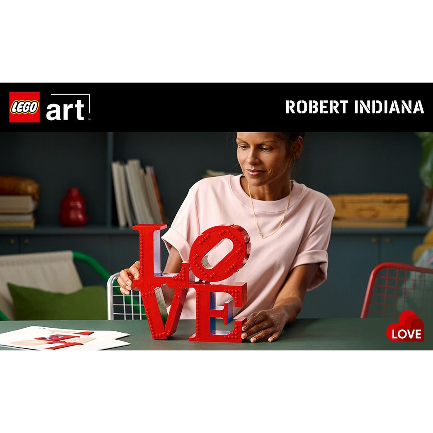 Donna con arte LEGO LOVE. Lettere rosse su un tavolo. Il logo LEGO è in alto a sinistra, con il nome di Robert Indiana.