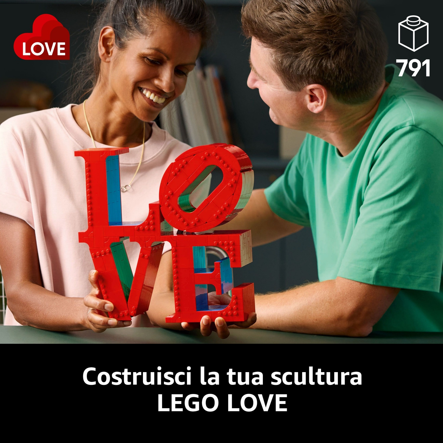 Una coppia sorride, tenendo una scultura LEGO rossa 'LOVE'. L'uomo veste di verde, la donna di rosa. Sfondo con libri e icona LEGO '791'.