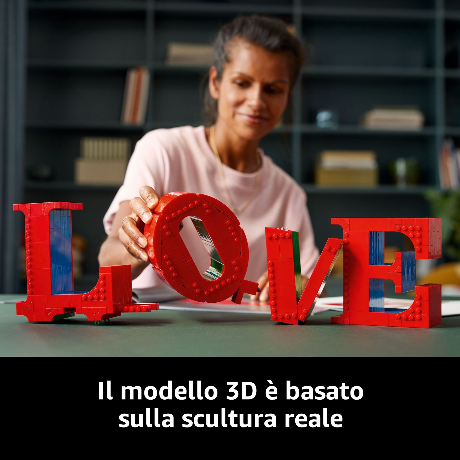 Una donna costruisce un cartello 'LOVE' LEGO rosso. La 'O' viene posizionata. Lo sfondo mostra una libreria con libri e un tavolo verde.