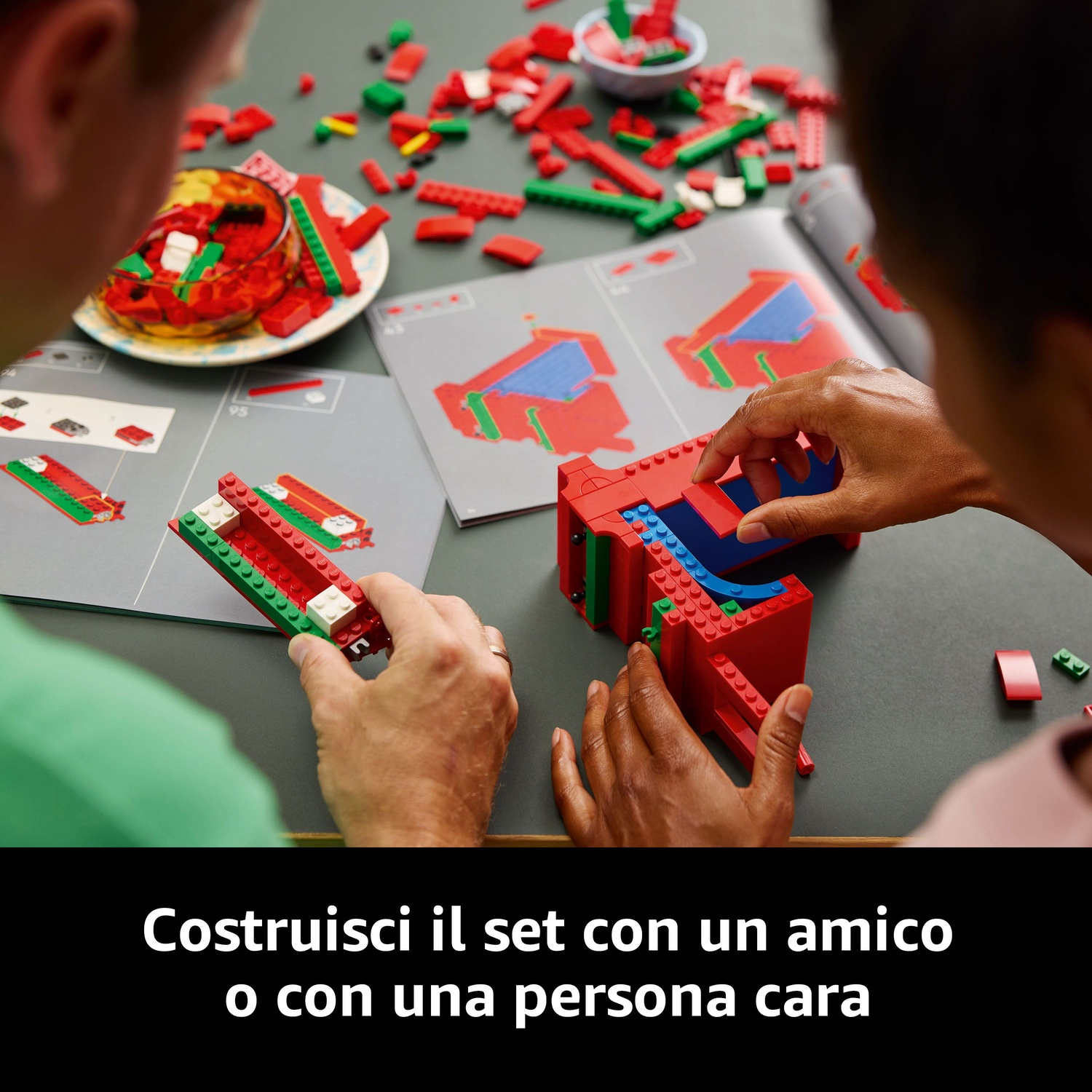 Due persone che costruiscono un set Lego. Mattoncini Lego rossi, verdi e blu su una superficie grigia con istruzioni e una ciotola.