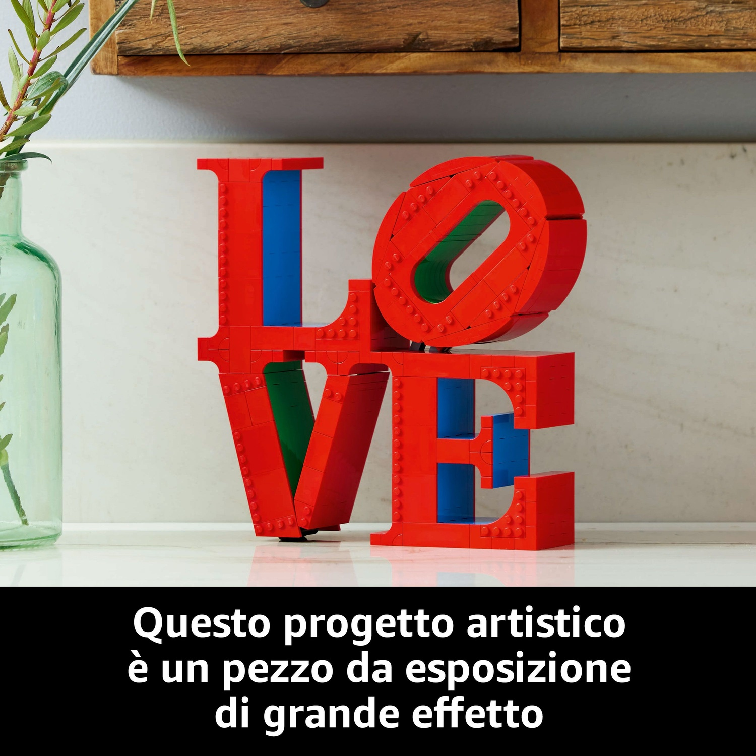 Scultura 'LOVE' rossa fatta di blocchi, con accenti blu e verdi, su una superficie bianca. Un vaso con piante verdi a sinistra.