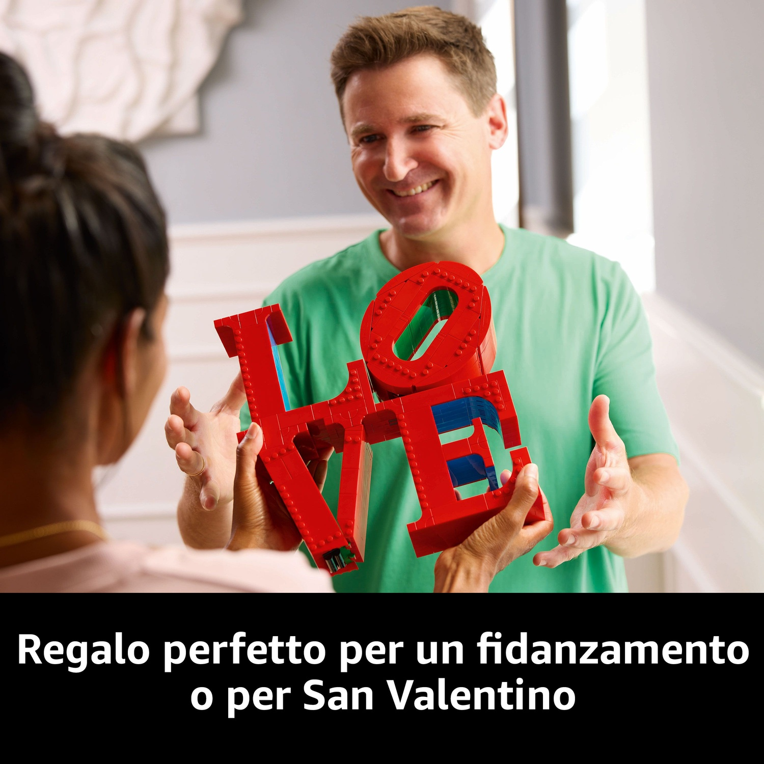Un uomo offre un cartello LEGO 'LOVE' rosso a una donna. Entrambi sorridono. Il testo sottostante dice: 'Regalo perfetto per un fidanzamento o San Valentino'.