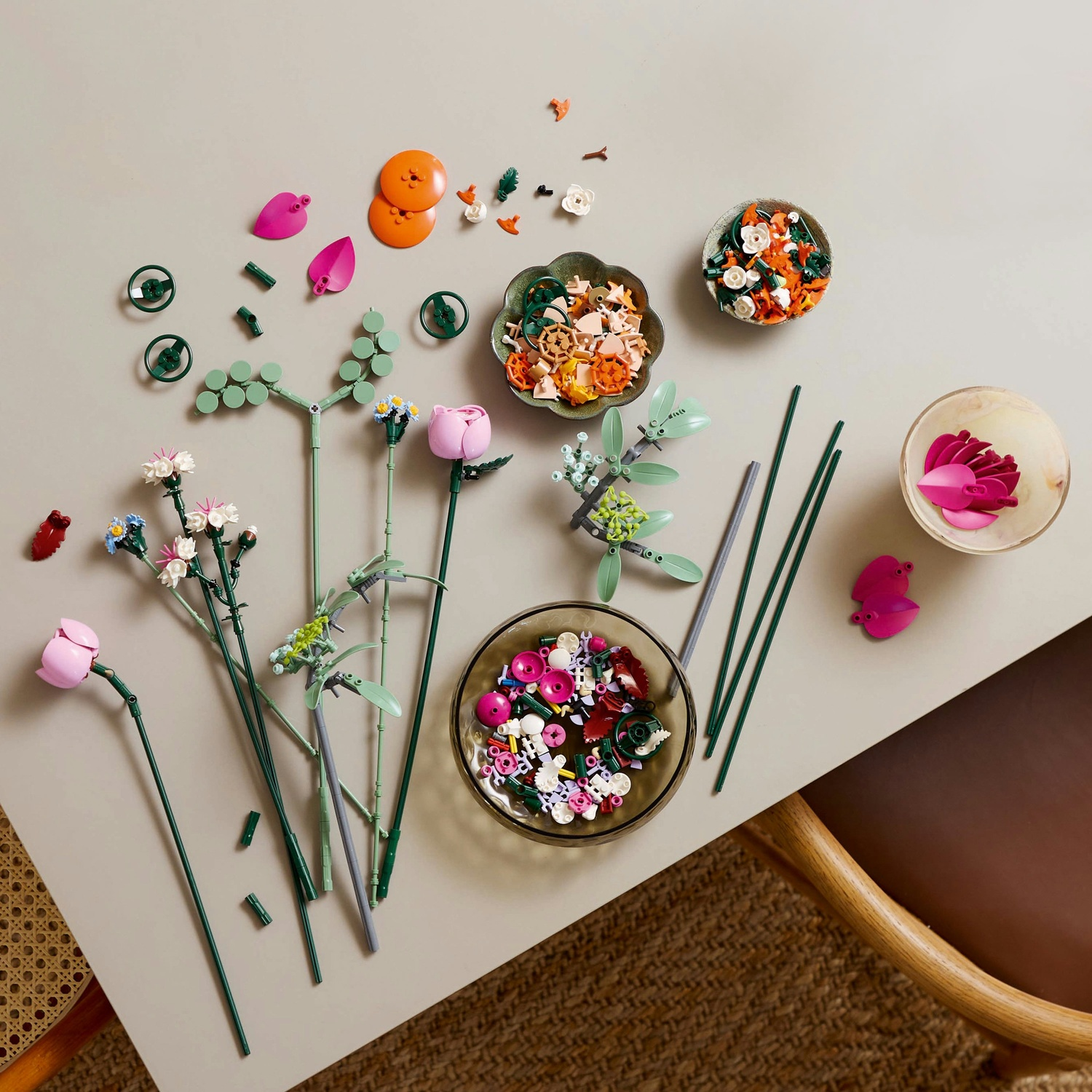 Vista dall'alto di pezzi di fiori Lego, tra cui petali rosa e arancioni, steli di fiori e ciotole di pezzi assortiti su un tavolo.